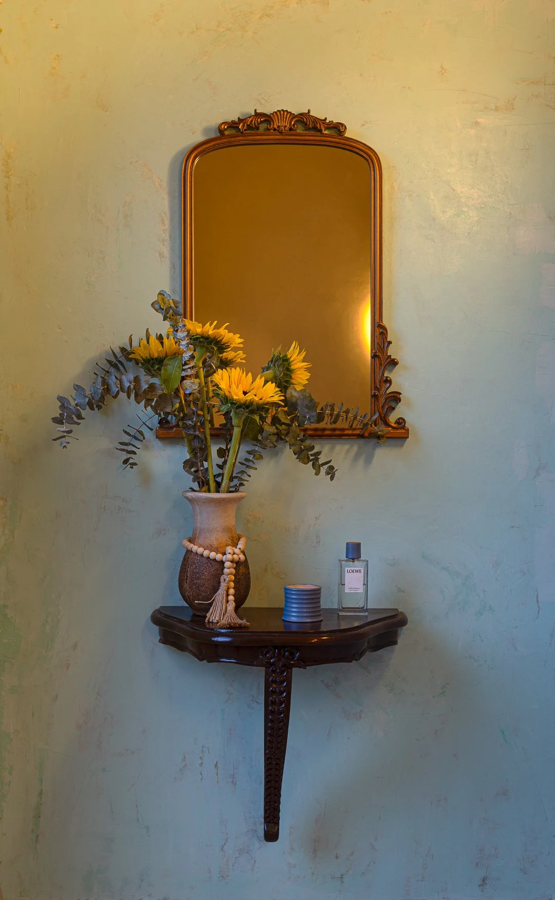 A decorative wall mirror with a wooden frame hanging above a small ornate shelf. On the shelf, a flower vase with sunflowers and greenery, a candle, and a bottle of perfume are displayed.