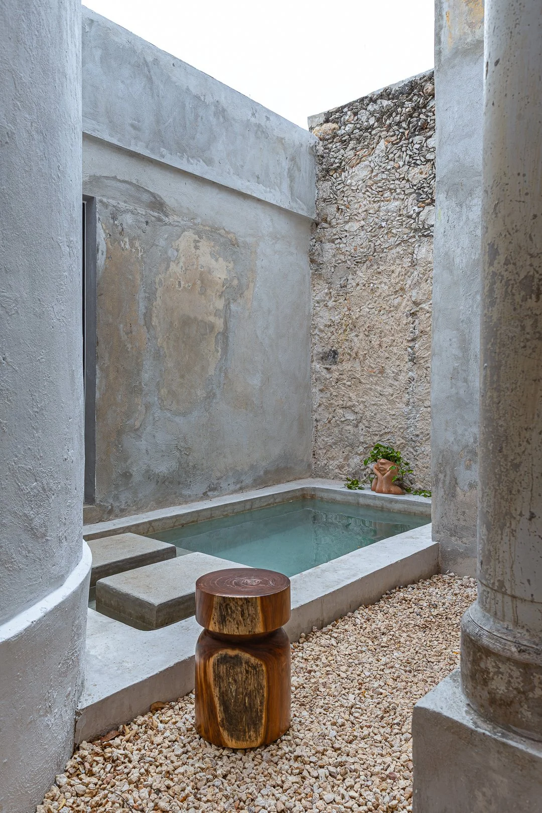 A small outdoor pool surrounded by stone and concrete walls, with a rustic wooden stool and a small statue of a woman with plants on her head at one corner.