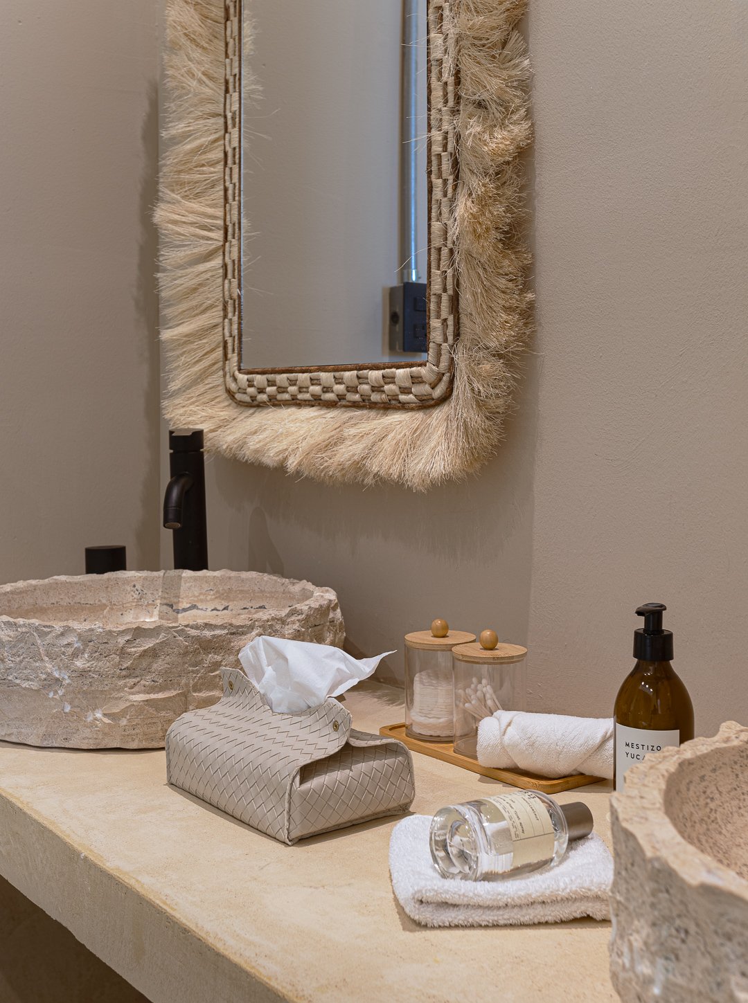 Bathroom countertop with stone sink, tissue box, glass bottles with lids, rolled towel, soap dispenser, and a mirror with a woven fur frame