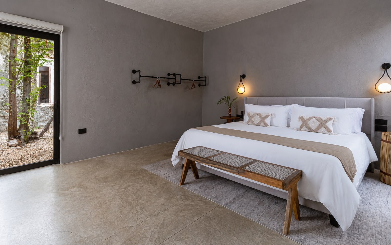 A modern bedroom with a large bed with white linens, decorative pillows, and a beige runner. There are two wall-mounted lamps above the bed, and a small side table with a plant next to it. A wooden bench is placed at the foot of the bed. A sliding gl