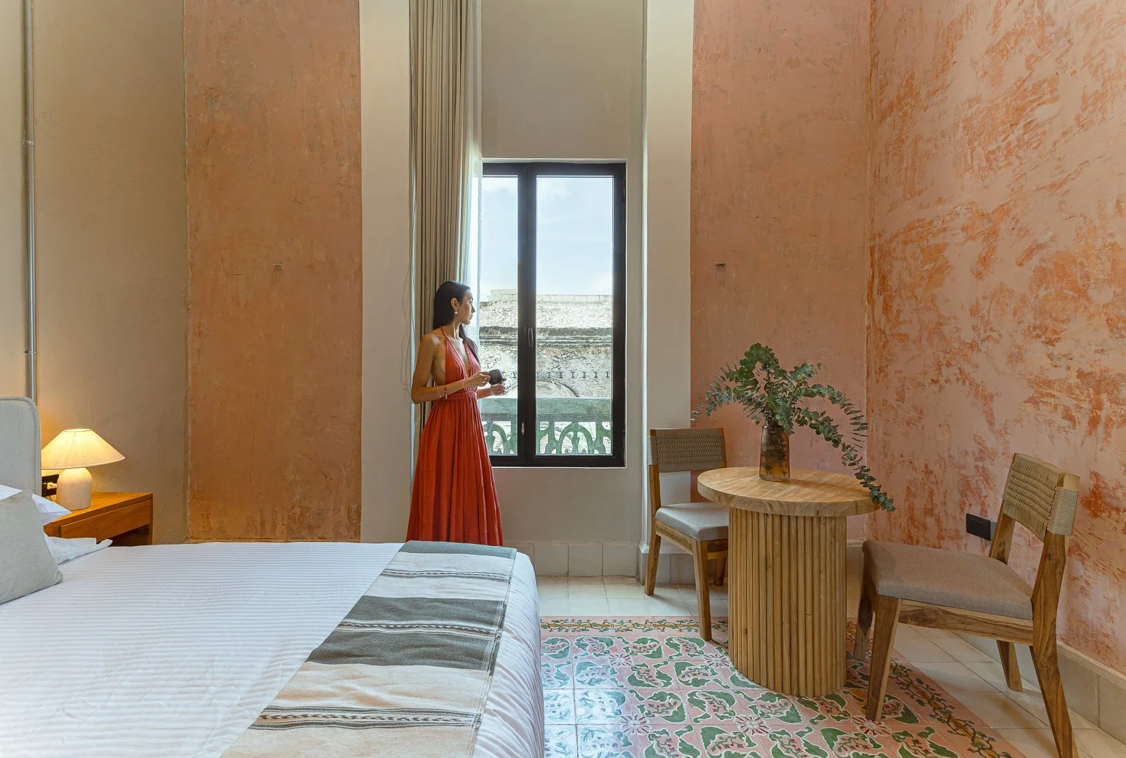 A woman in a red dress standing by a large window in a warmly lit bedroom with textured pink walls, a bed with white linens, a bedside table with a lamp, a round wooden table with a vase of green foliage, and two chairs.
