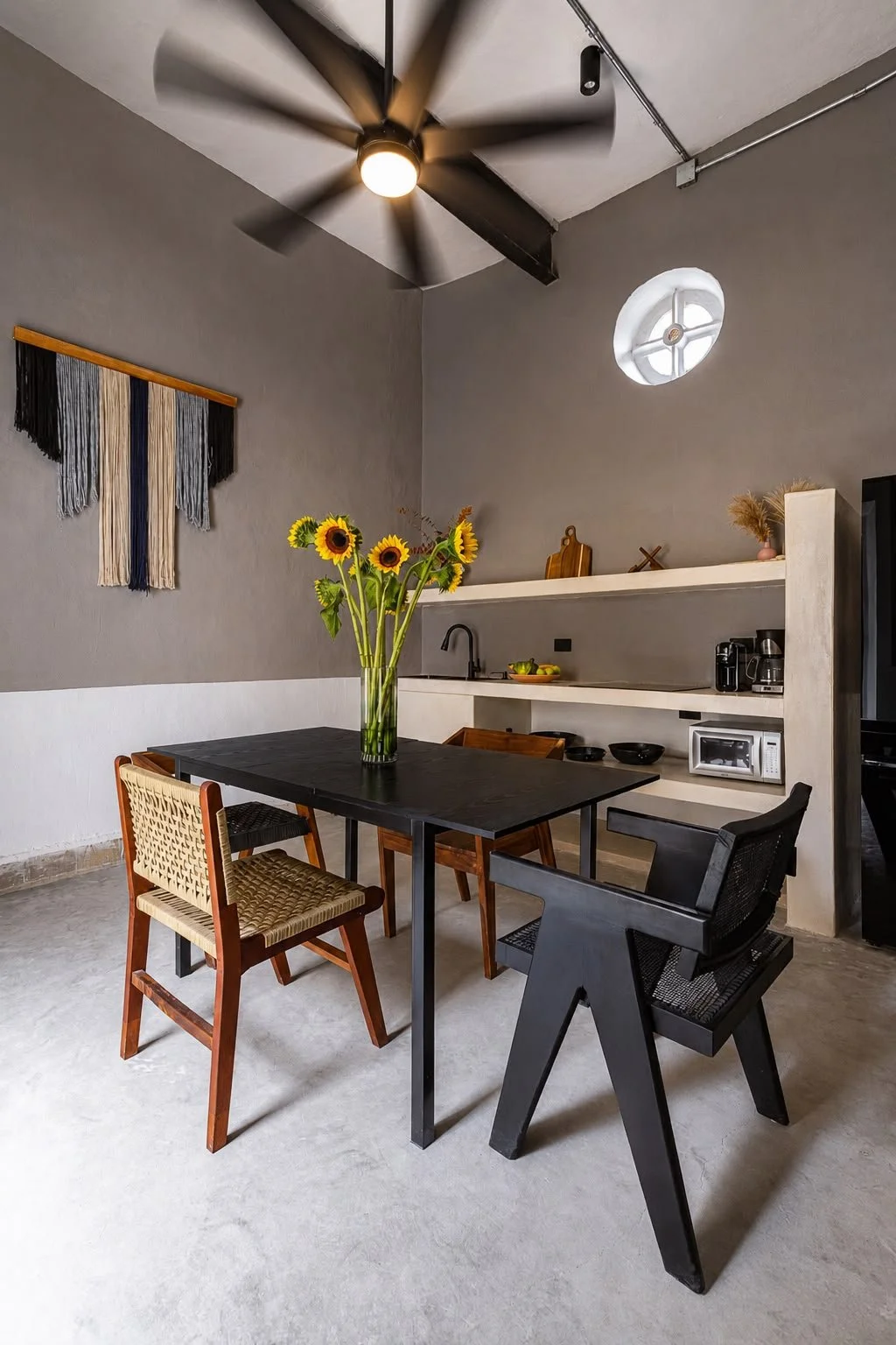 Modern dining area with a black table, a vase of sunflowers, and mixed chairs, with a ceiling fan and wall decor.