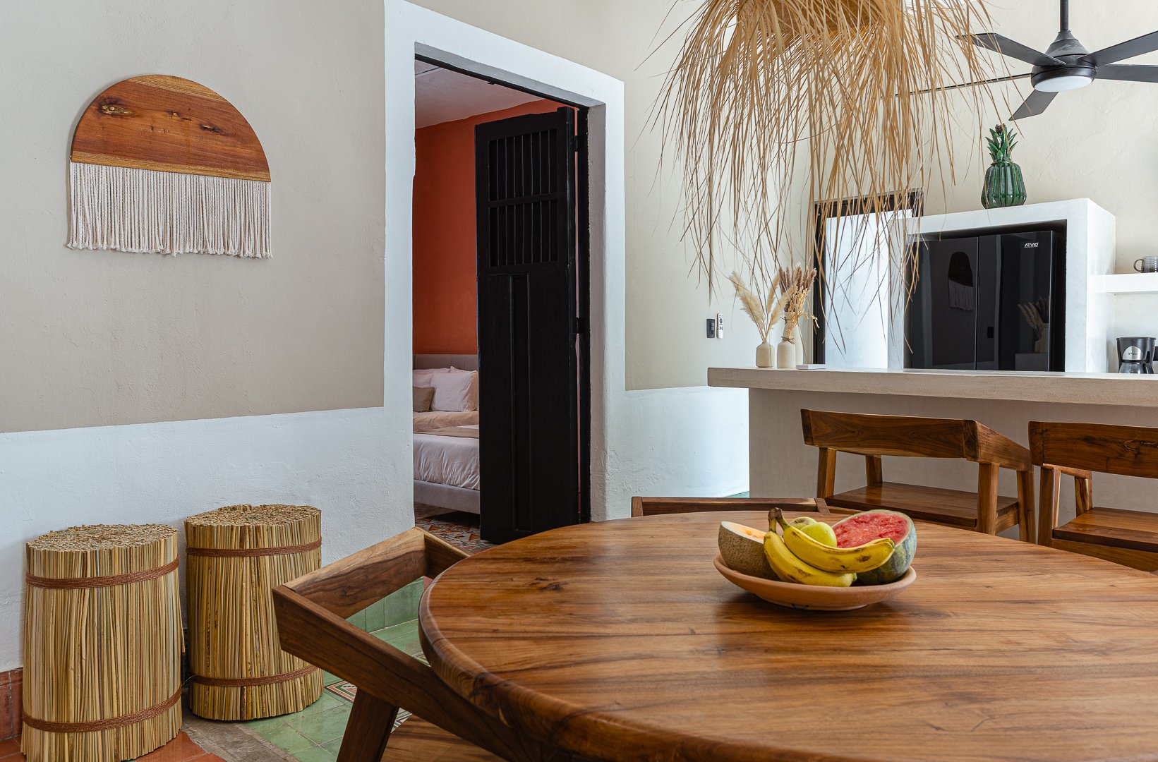 A cozy dining area with a round wooden table, a bowl of mixed fruit, and wooden chairs. In the background, there is a kitchen counter with decor vases and a ceiling fan, and a doorway leading to a bedroom with a partially visible bed.