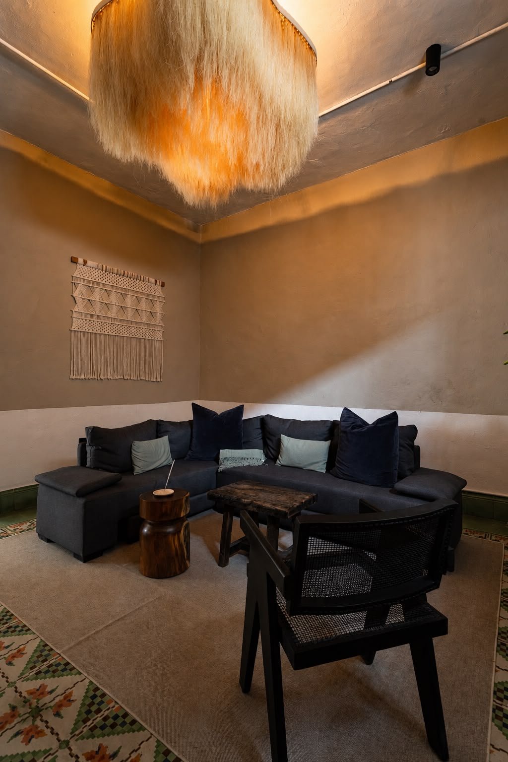 A cozy living room with a dark gray sectional sofa with throw pillows, a small wooden side table, a black chair, a wooden coffee table, a beige rug, and a macrame wall hanging, illuminated by a large orange furry ceiling light.