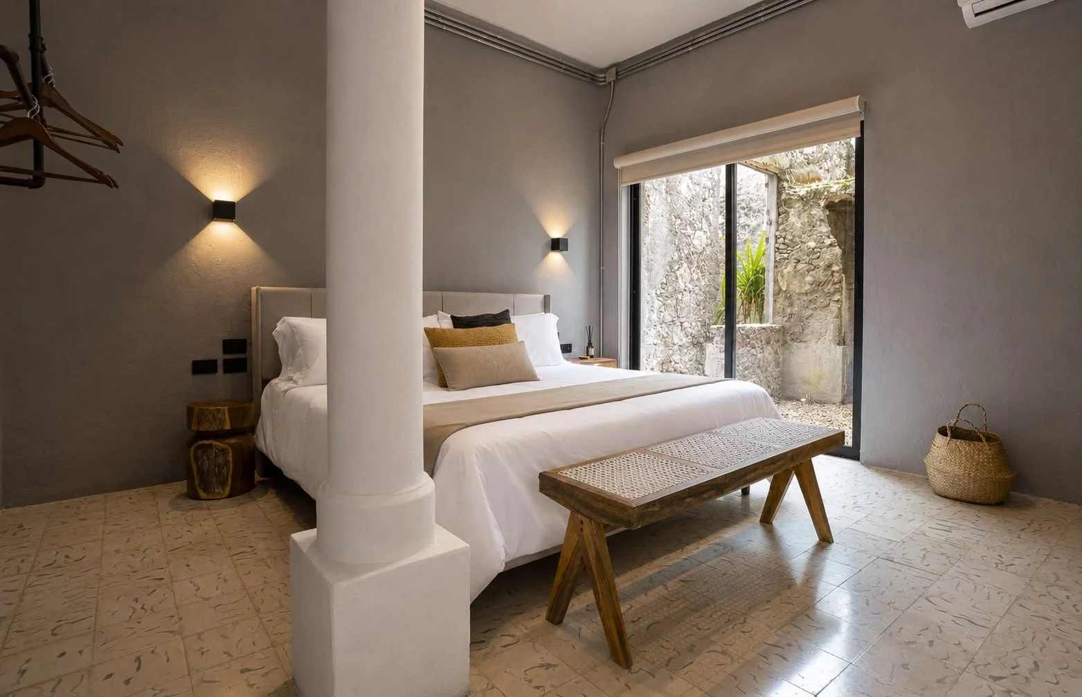 A bedroom with a large bed, beige bedding, and decorative pillows. There's a wooden bench at the foot of the bed, a rustic side table, a basket, and sliding glass doors leading to an outdoor stone patio with plants.