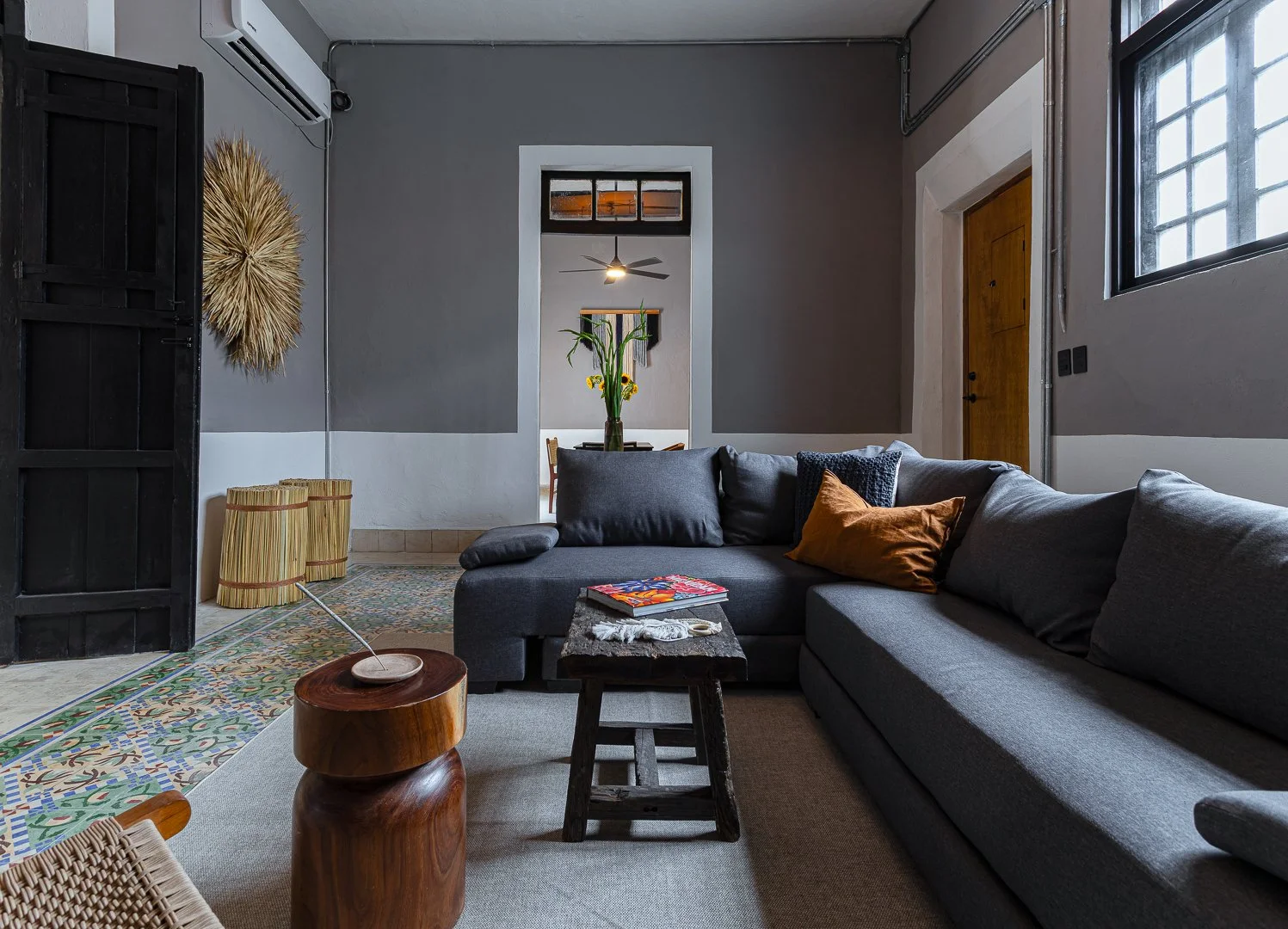 Living room with gray sectional sofa with colorful pillows, black wooden coffee table, patterned rug, and decorative wall art, with a view into a dining room with a tall vase of green plants and sunflowers.