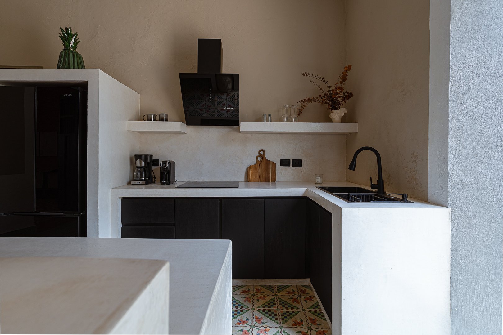 A minimalist kitchen with black lower cabinets, a white countertop, and a black sink with a curved faucet. White floating shelves hold glassware, cups, and a decorative plant. A built-in black range hood is above the stovetop, with a cutting board an
