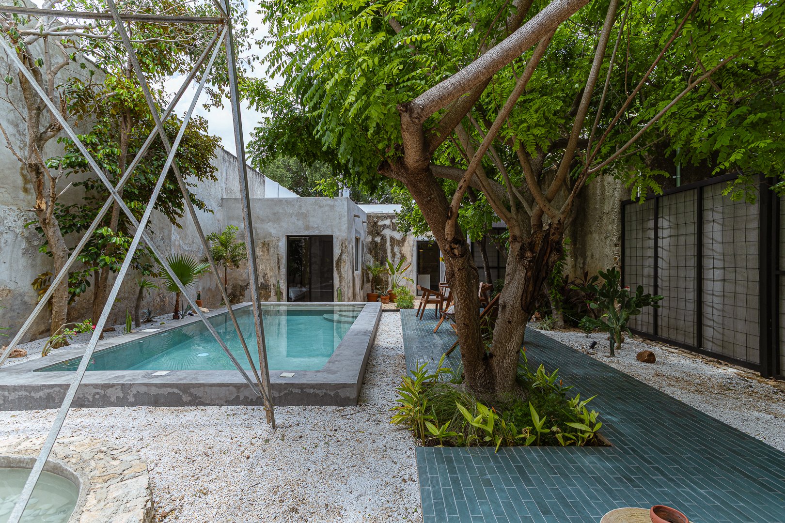 Urban backyard with a small rectangular pool, surrounded by gravel, lush green trees, and plants, with modern concrete walls and a seating area with wooden chairs.