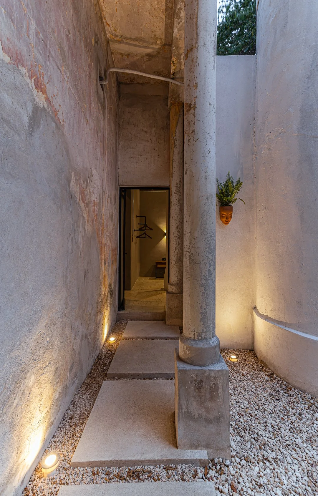 Exterior view of a minimalist courtyard with concrete flooring, a carved stone pillar, a wall-mounted potted plant, and a doorway leading to an interior space with wooden coat hangers.
