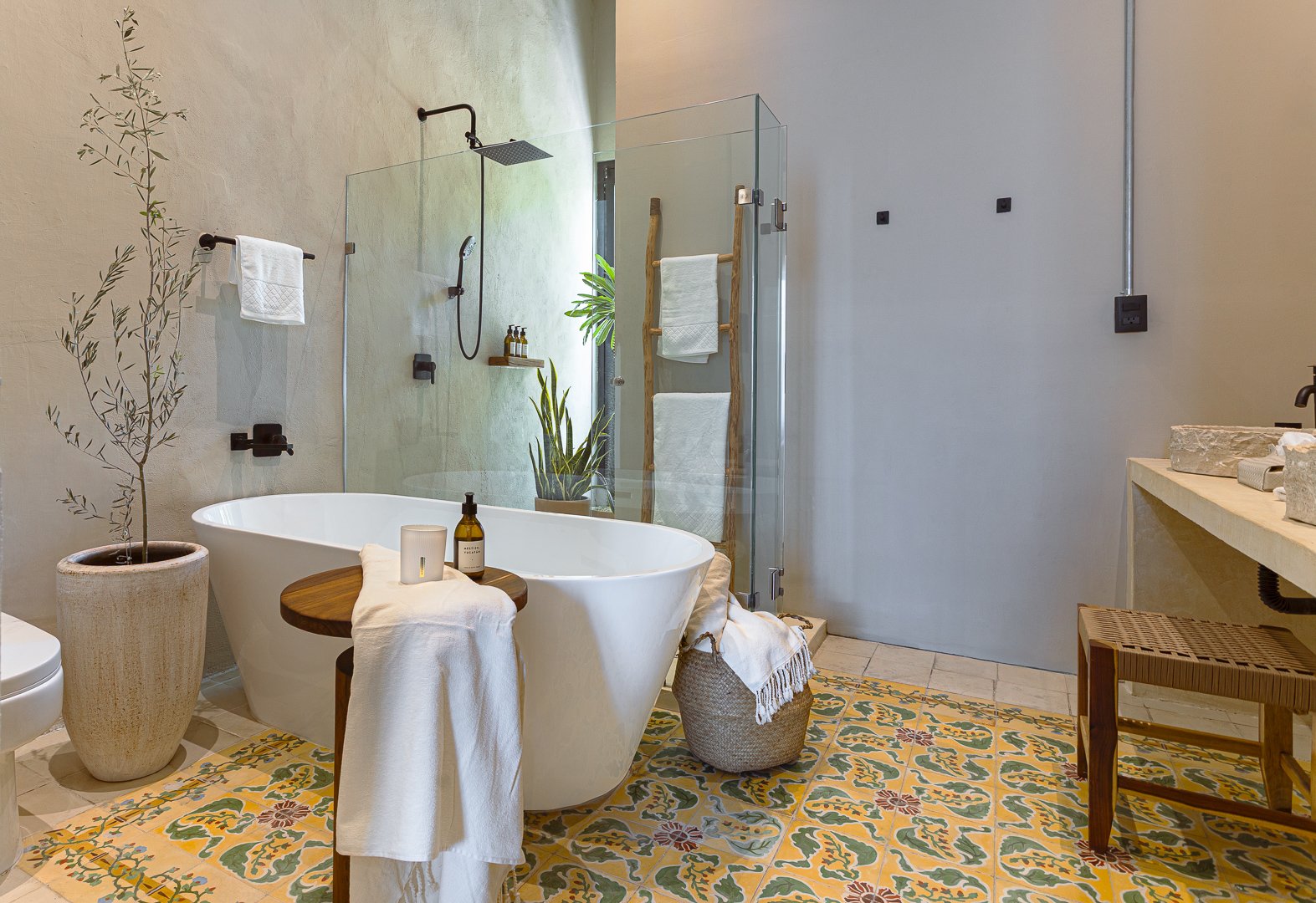 Modern bathroom featuring a freestanding bathtub with wooden side table and towel, potted trees, a glass-enclosed shower, a patterned tile floor, and minimalist fixtures.
