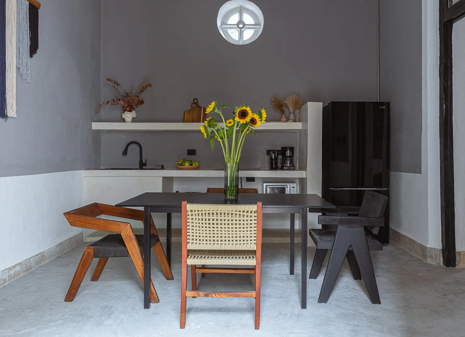 Modern dining room with a black table, four chairs of different designs, and a sunflower centerpiece in a glass vase. In the background, a kitchenette with a black sink, coffee maker, microwave, and decorative items on a shelf.