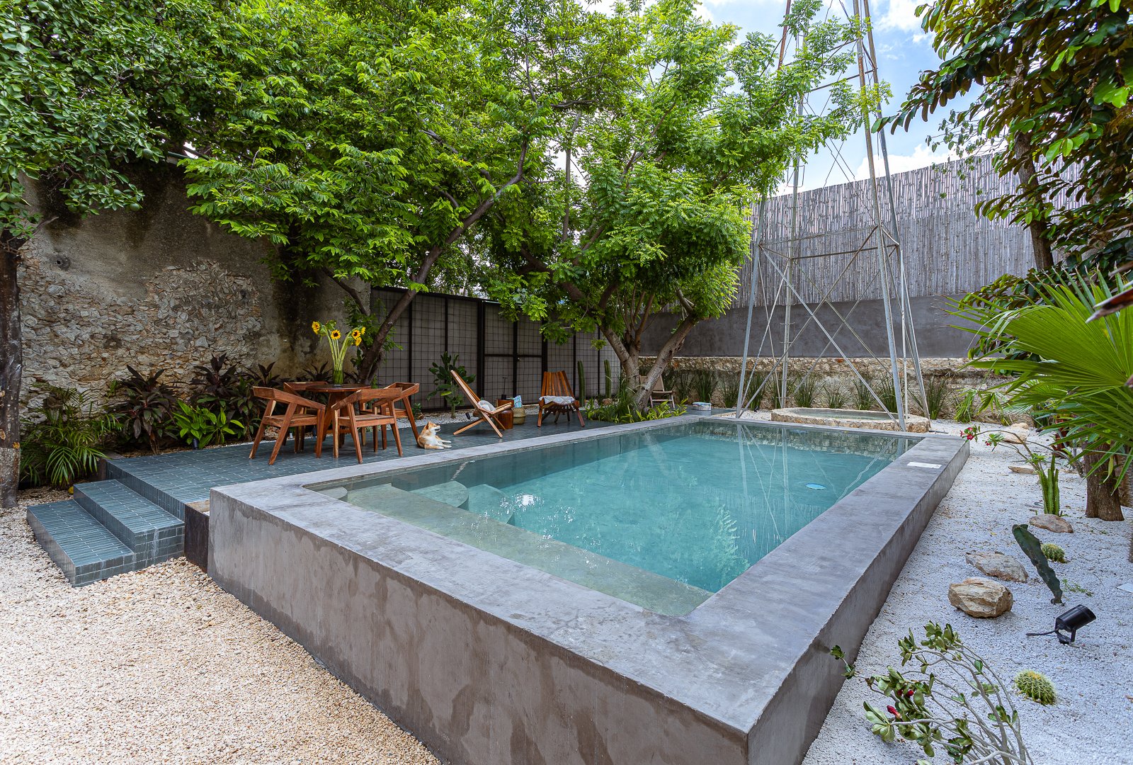 A backyard with a small pool, outdoor deck with chairs, trees, plants, and a windmill structure.