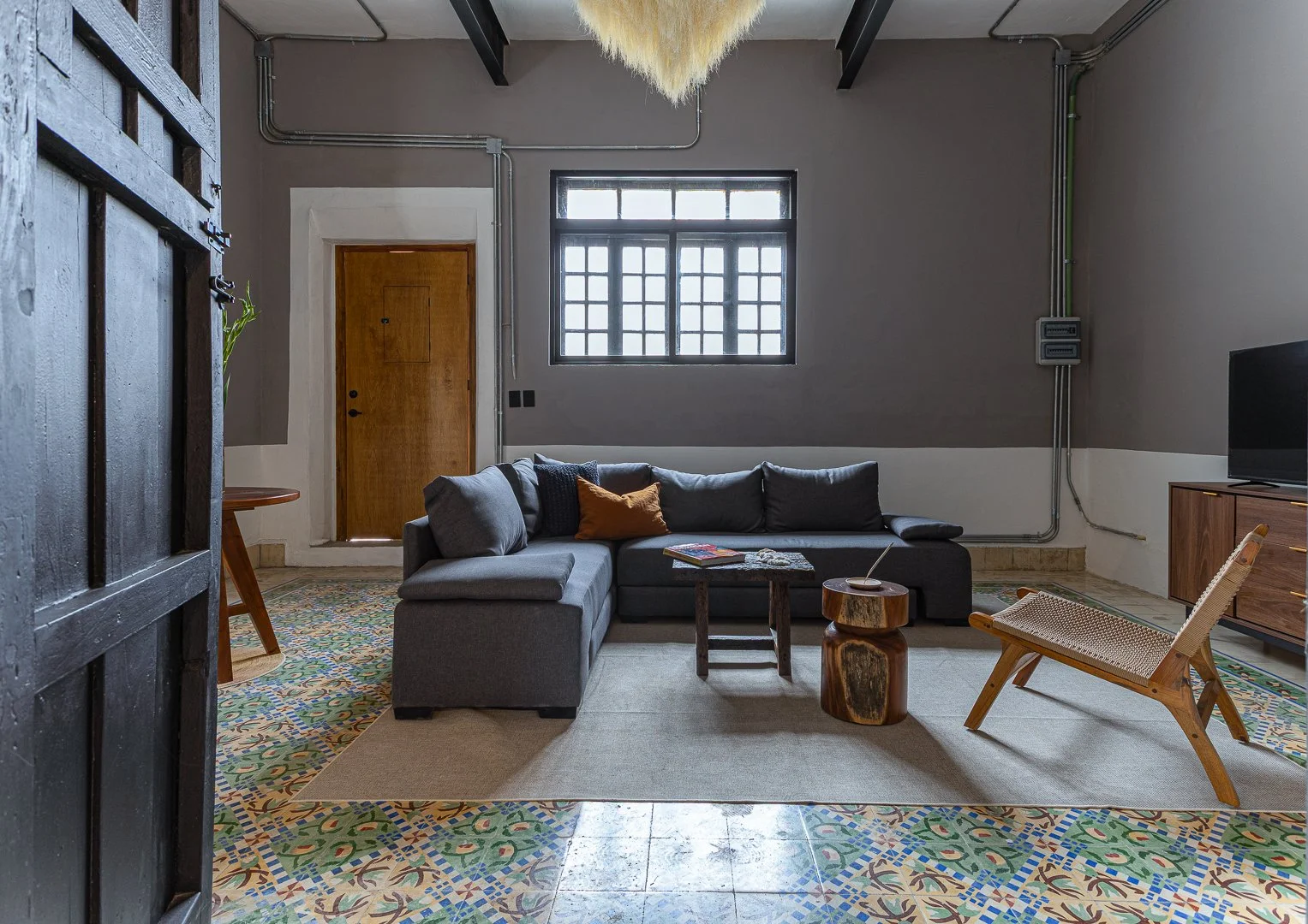 Living room with gray sectional sofa, wooden side table, colorful patterned tile floor, and a window on the gray wall. There is a black sliding door partially open on the left, with a small table behind it. The room has a modern rustic decor style.