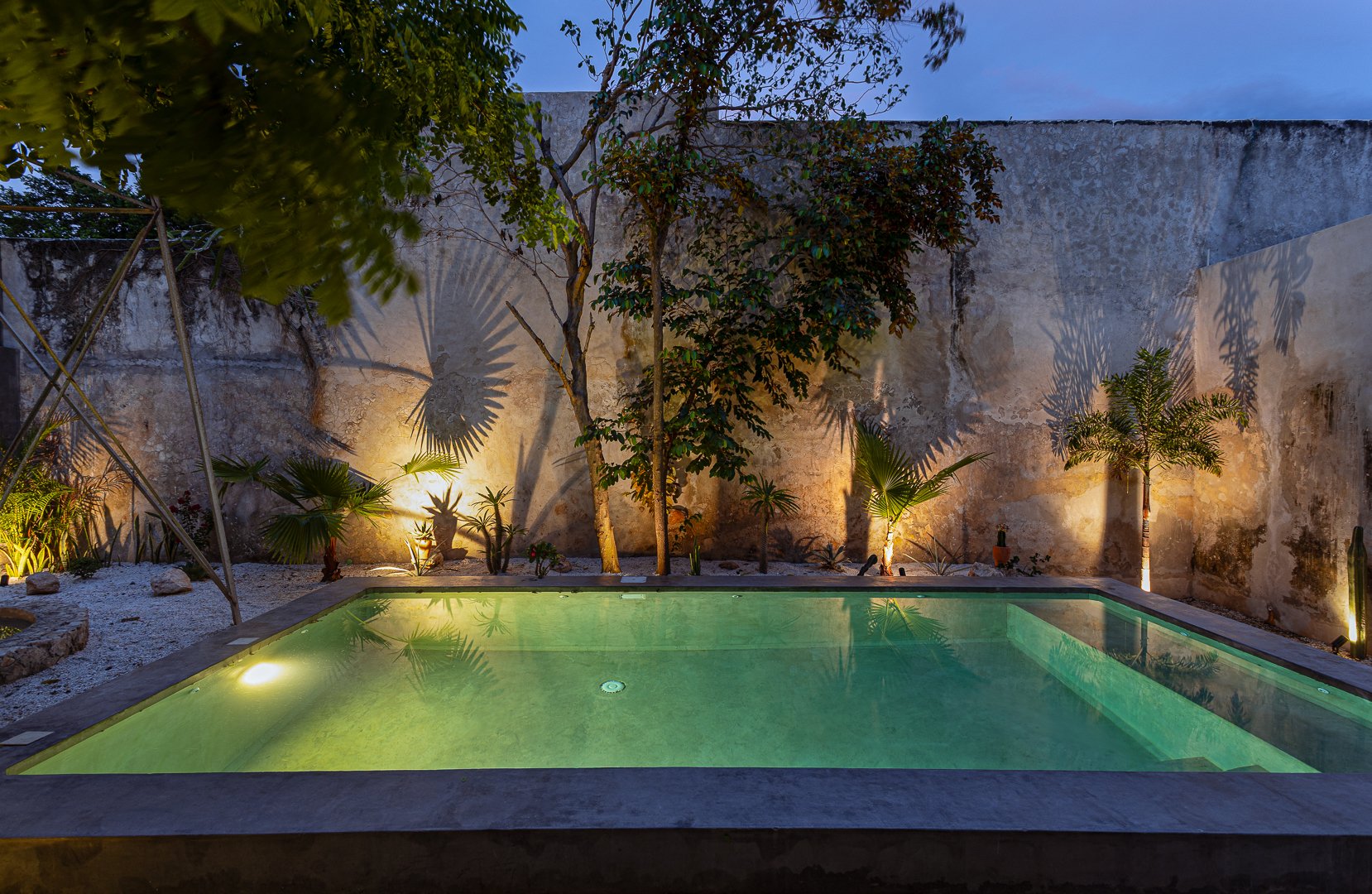 A small illuminated swimming pool in a courtyard with a stone wall, trees, and decorative plants, nighttime setting.