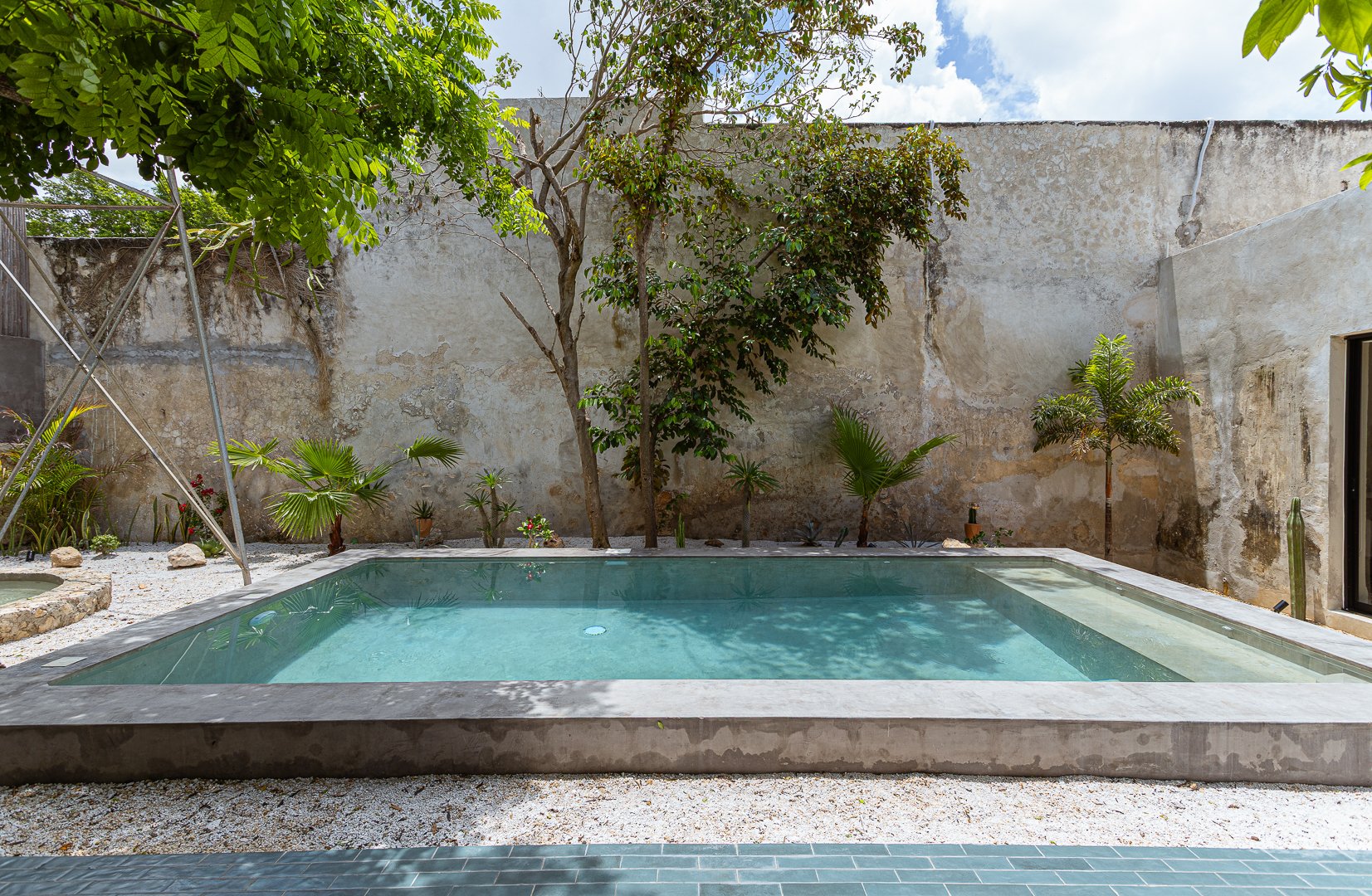 A small backyard pool surrounded by a concrete border, with a weathered concrete wall in the background and tropical plants, including palm trees, along the perimeter. A metallic structure is partially visible on the left, and the sky is partly cloud
