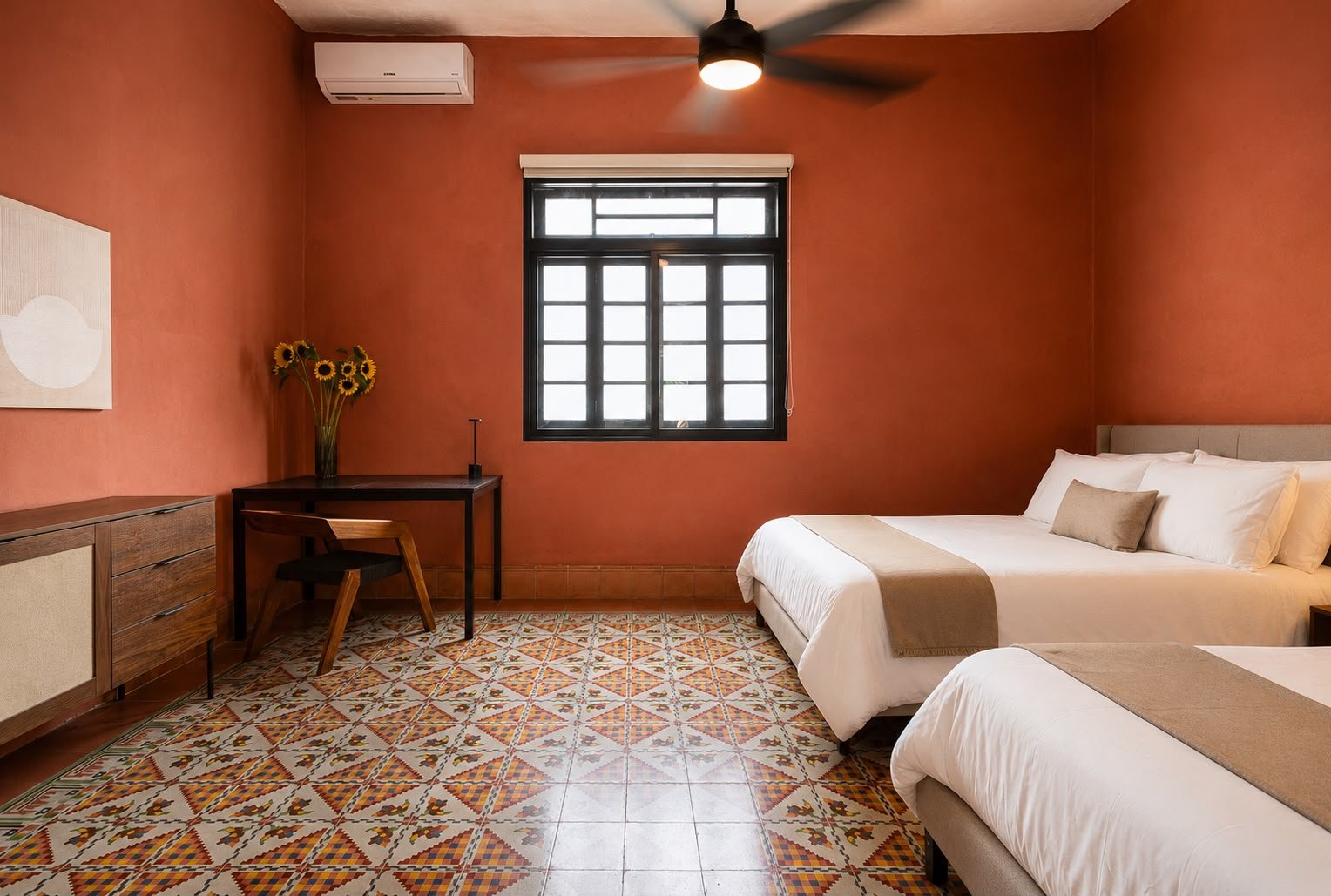 A hotel room with two beds, a black ceiling fan, a window with black framing, a wooden desk with a vase of sunflowers, a wooden chair, a cabinet, an air conditioning unit, and a patterned tiled floor, all with warm-colored walls.