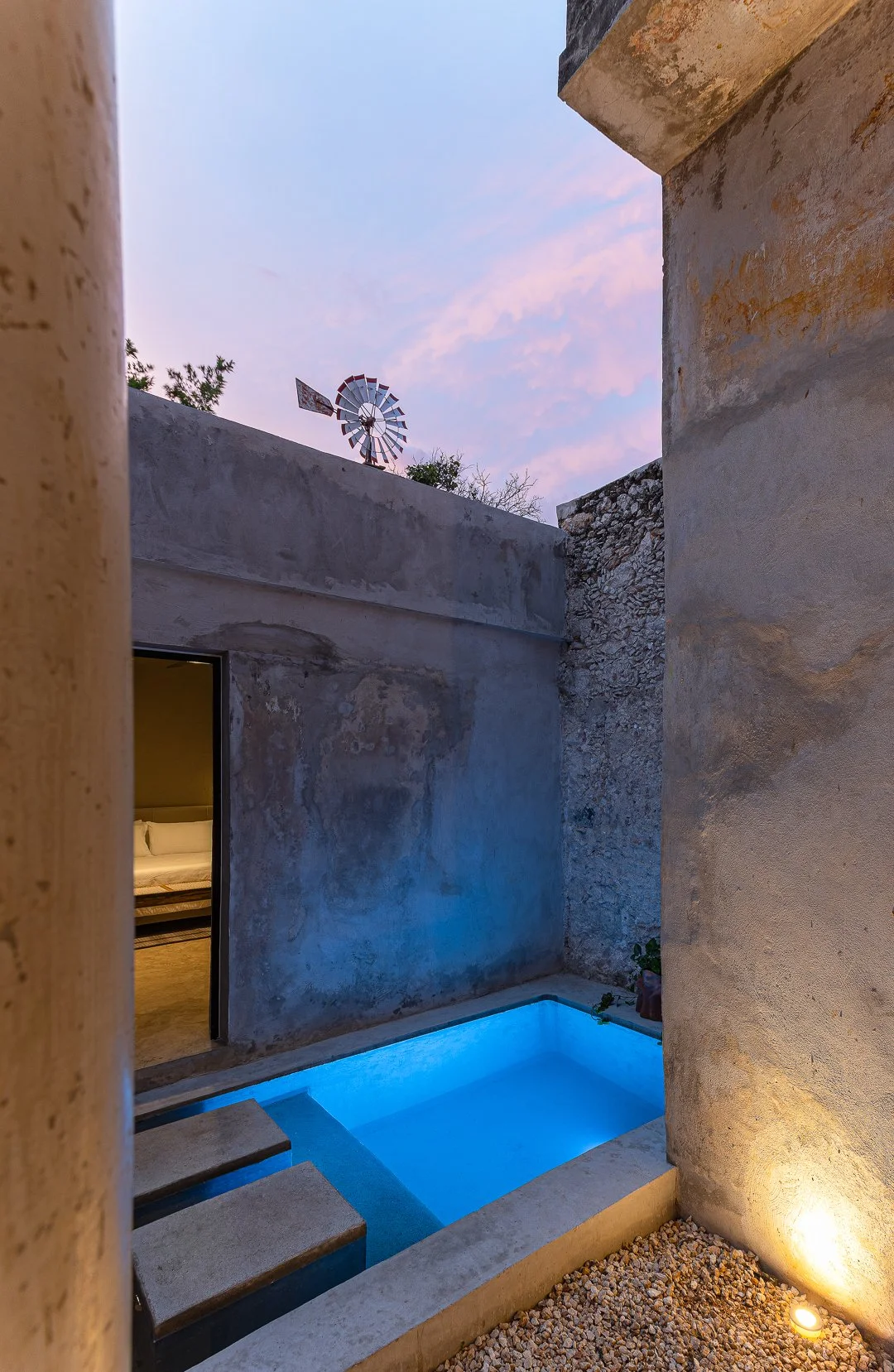 A small, illuminated blue pool situated outdoors between rustic concrete and stone walls at dusk, with an open door revealing a bedroom inside, and a windmill on the roof against a pink and purple sky.