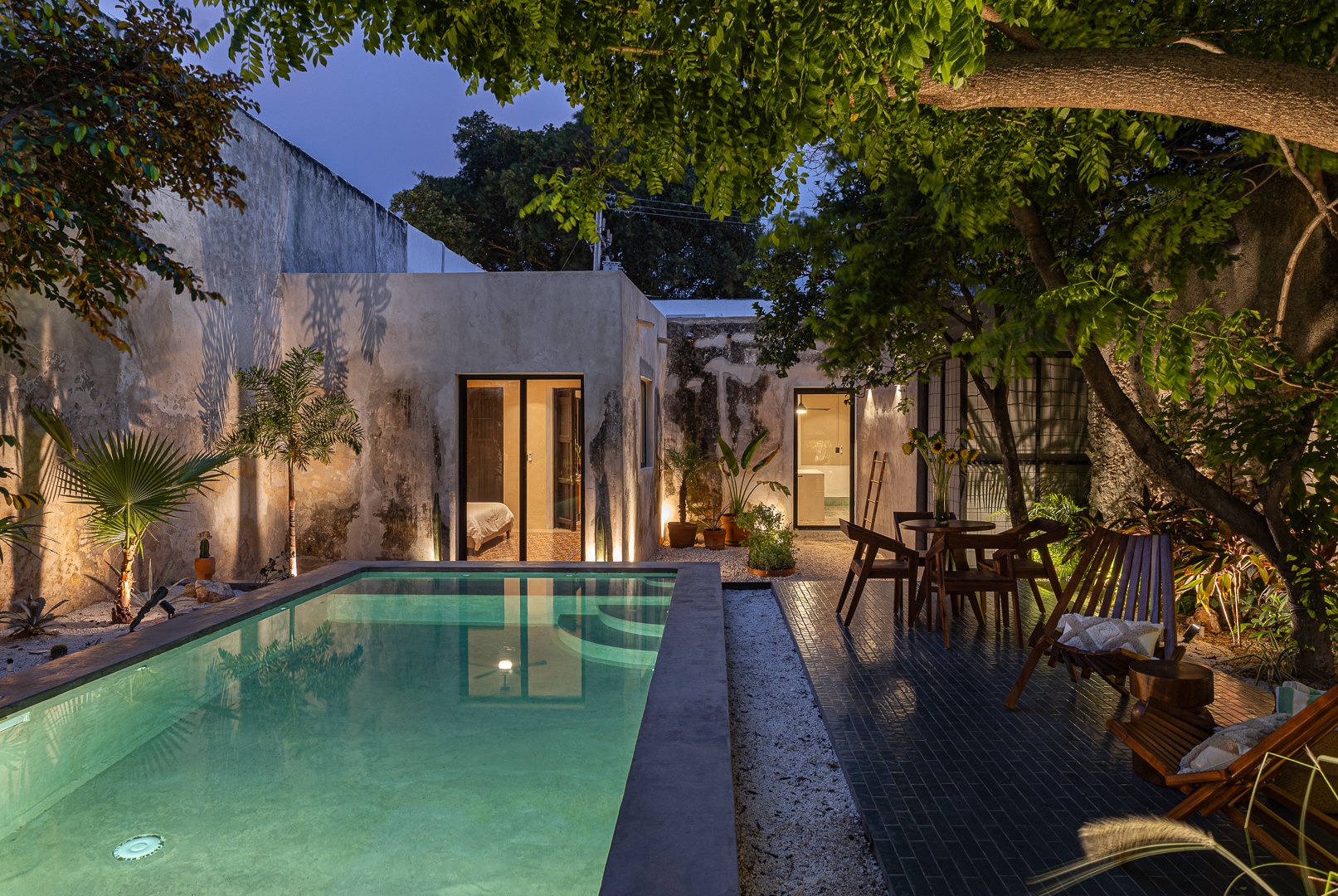 A cozy backyard with a small pool, surrounded by lush green plants and trees. There are outdoor chairs and a table on a dark tiled patio, with warm lighting illuminating the scene at dusk.