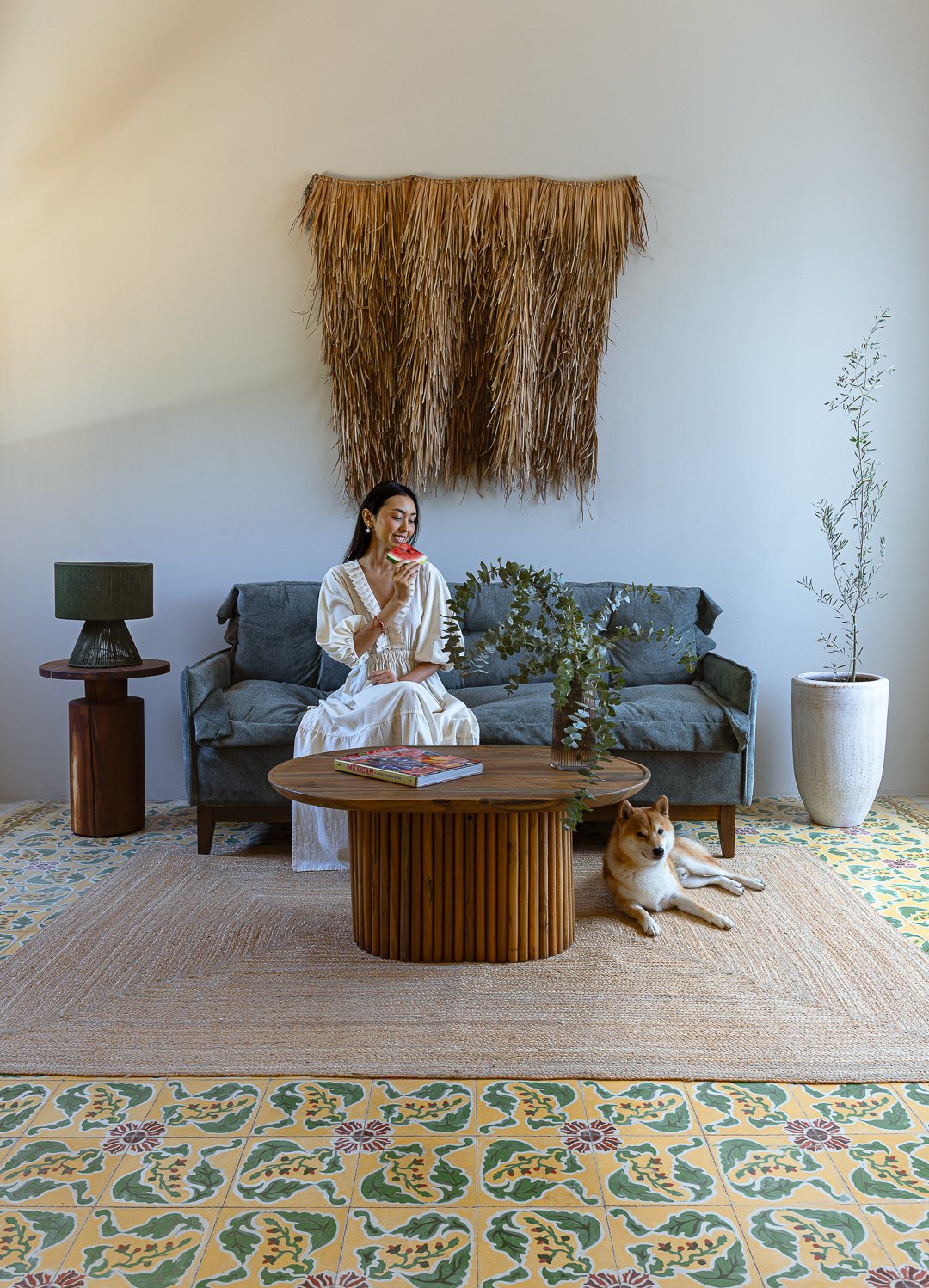 A woman in a beige dress sitting on a gray sofa with a dog lying on the patterned floor beside her. She is eating a slice of watermelon. There is a green plant on the coffee table, a dark green lampshade on a wooden side table, and two large potted p