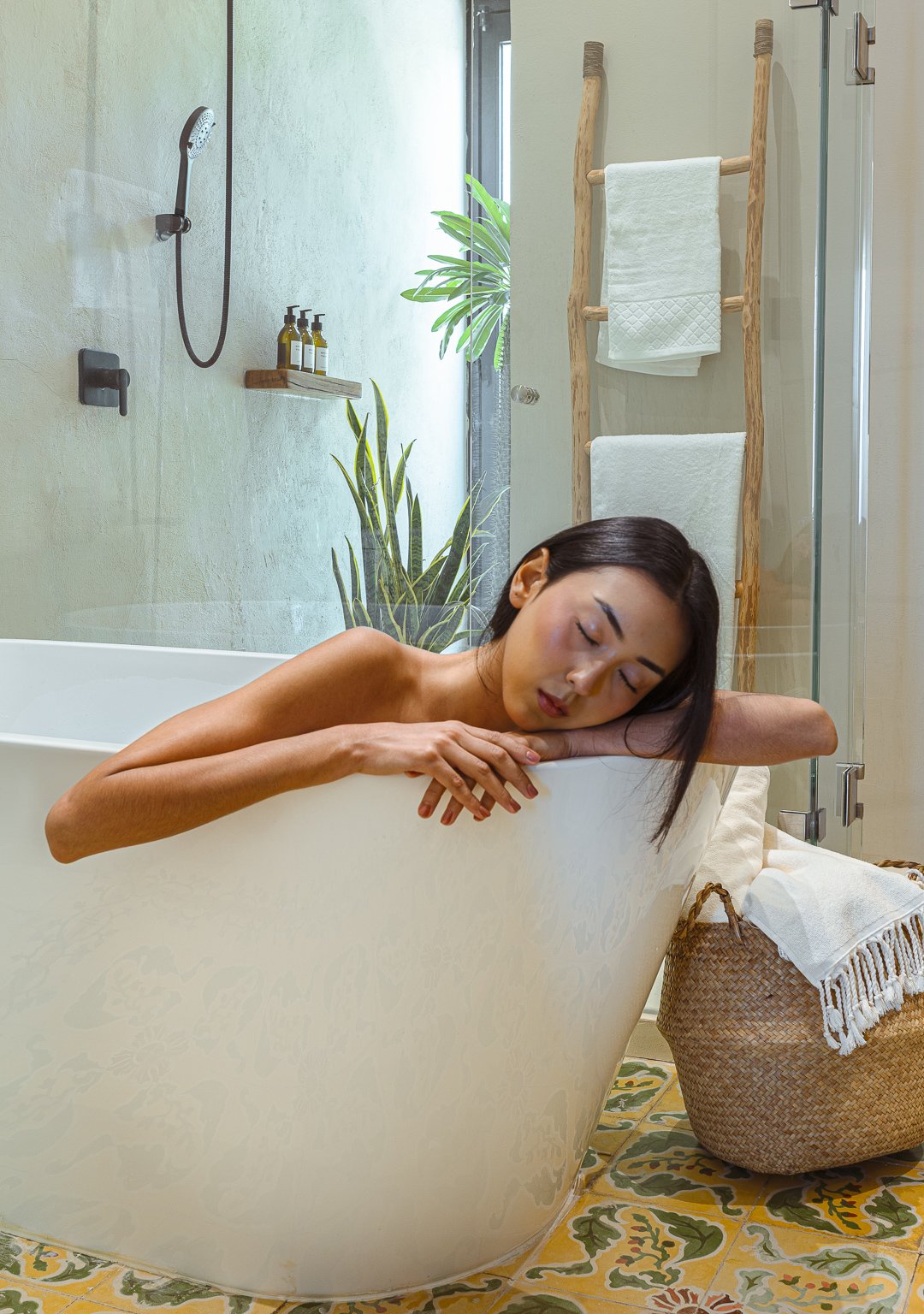 Woman relaxing in a bathtub in a bathroom with green walls, potted plants, wooden ladder towel rack, and patterned tiled floor.