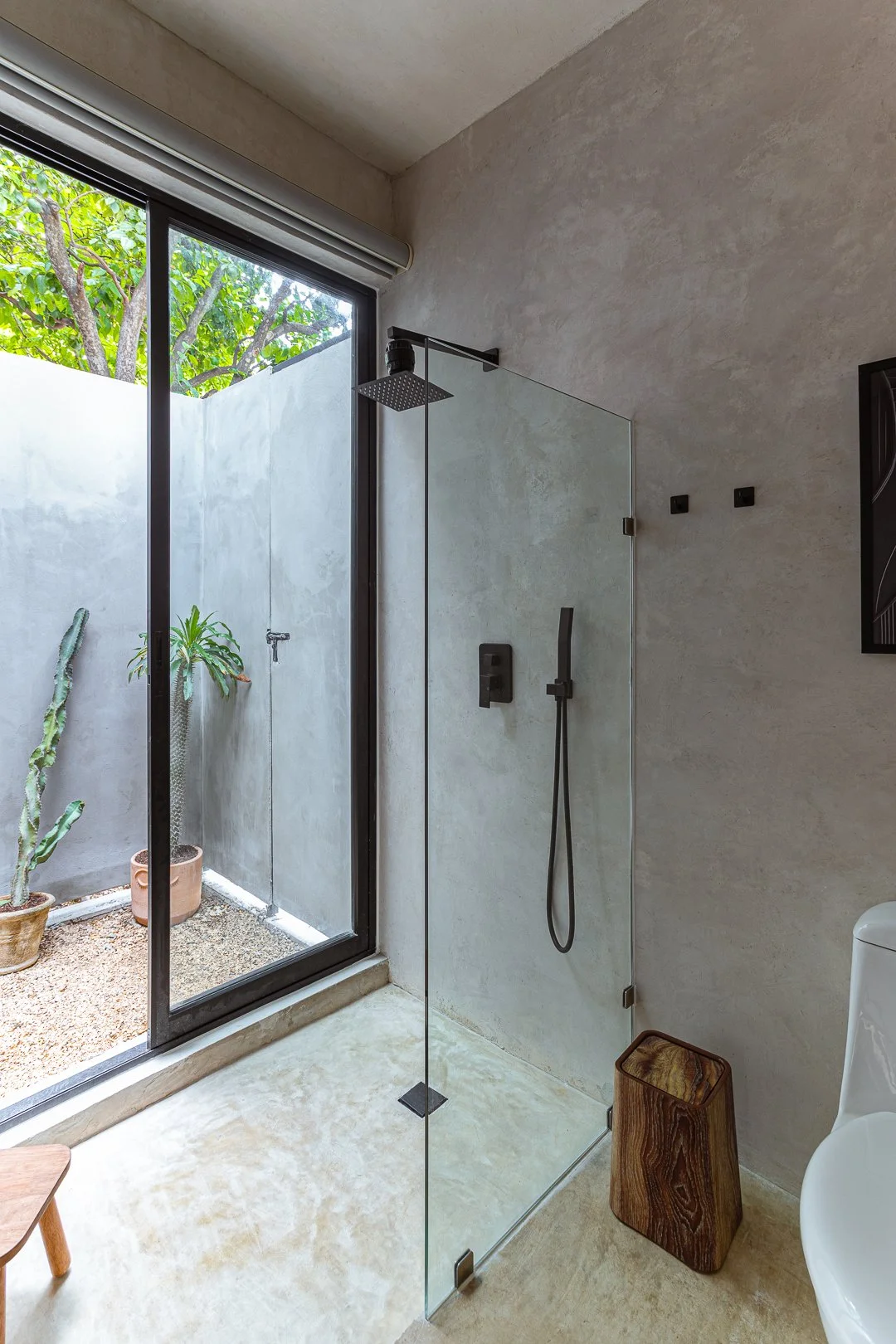 Modern minimalist bathroom with a walk-in shower featuring a black rain showerhead and black fixtures, adjacent to a large glass sliding door leading to an outdoor patio with potted plants, concrete walls, and a small wooden stool.