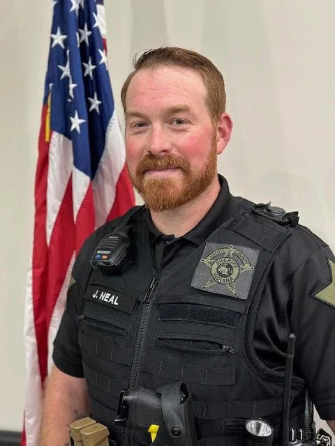 A police officer with a red beard and mustache standing in front of an American flag, wearing a black uniform with a badge and radio.