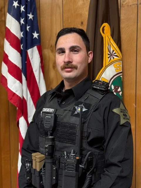 A male police officer stands indoors in front of an American flag and a department flag. He wears a black uniform with a vest and equipment, and has short dark hair and a mustache.