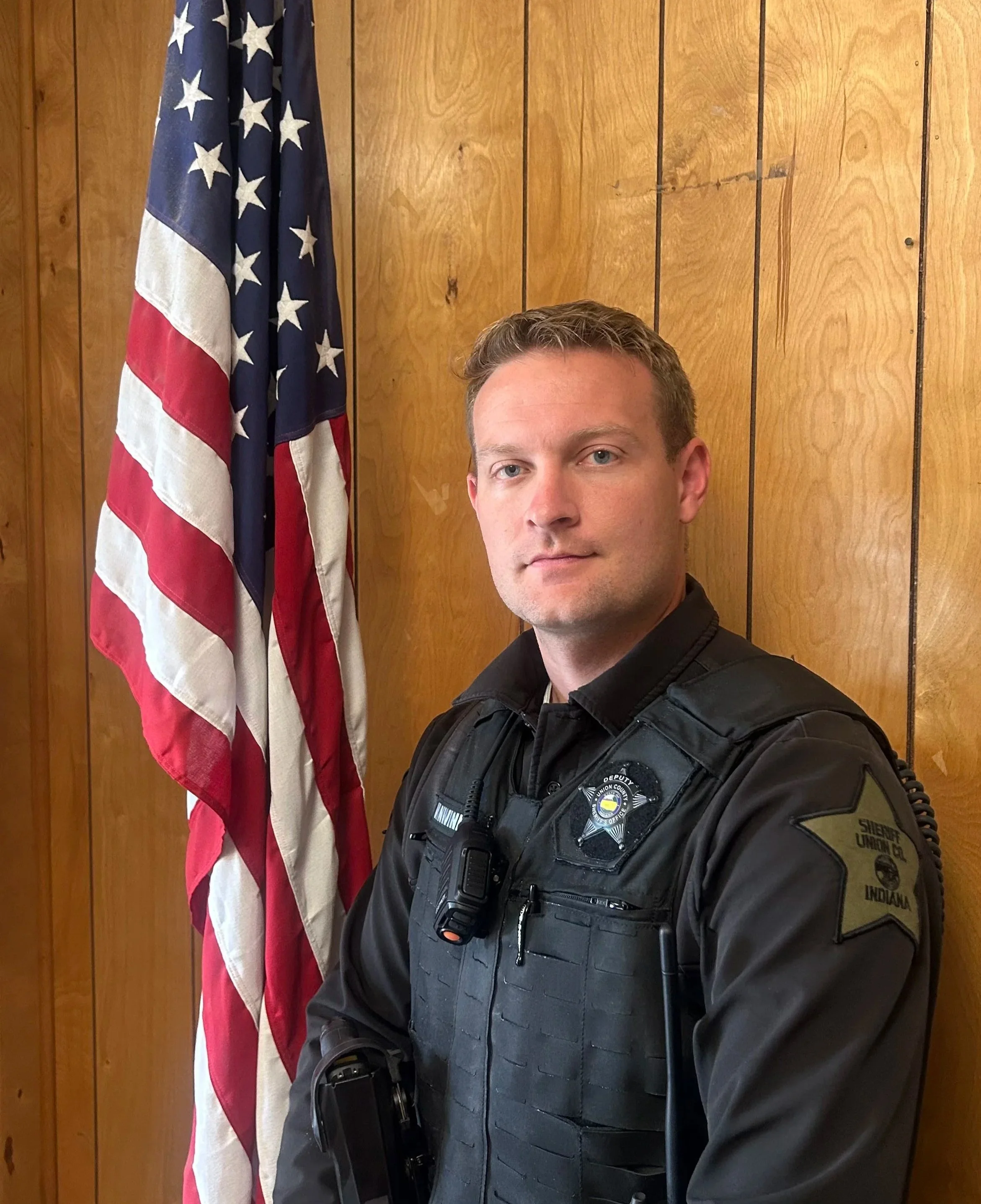 A man in a police uniform standing beside an American flag in front of a wooden wall.