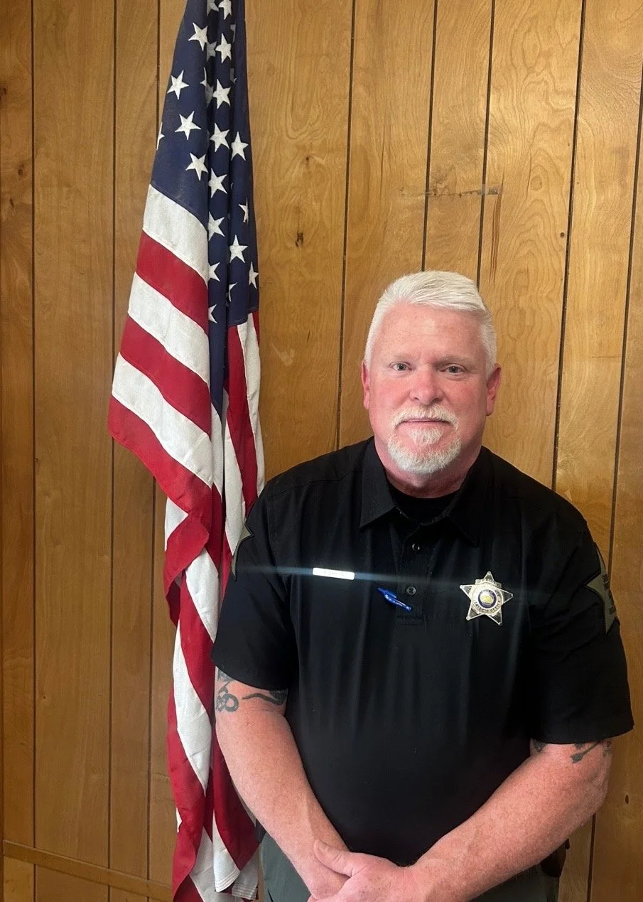 A man with white hair and a beard, wearing a black police uniform, standing in front of a wooden wall with an American flag to his left.