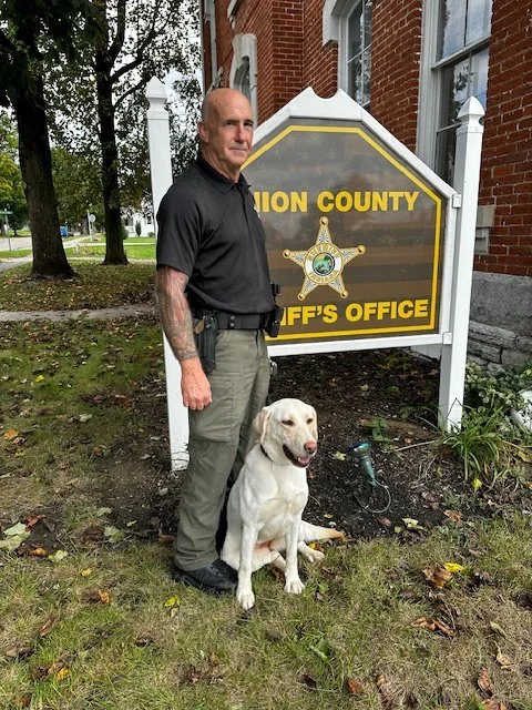 A man and a service dog standing outside a Union County Sheriff's Office sign. The man is bald, wearing a black polo shirt and khaki pants. The yellow Labrador Retriever service dog sits on the ground beside him.