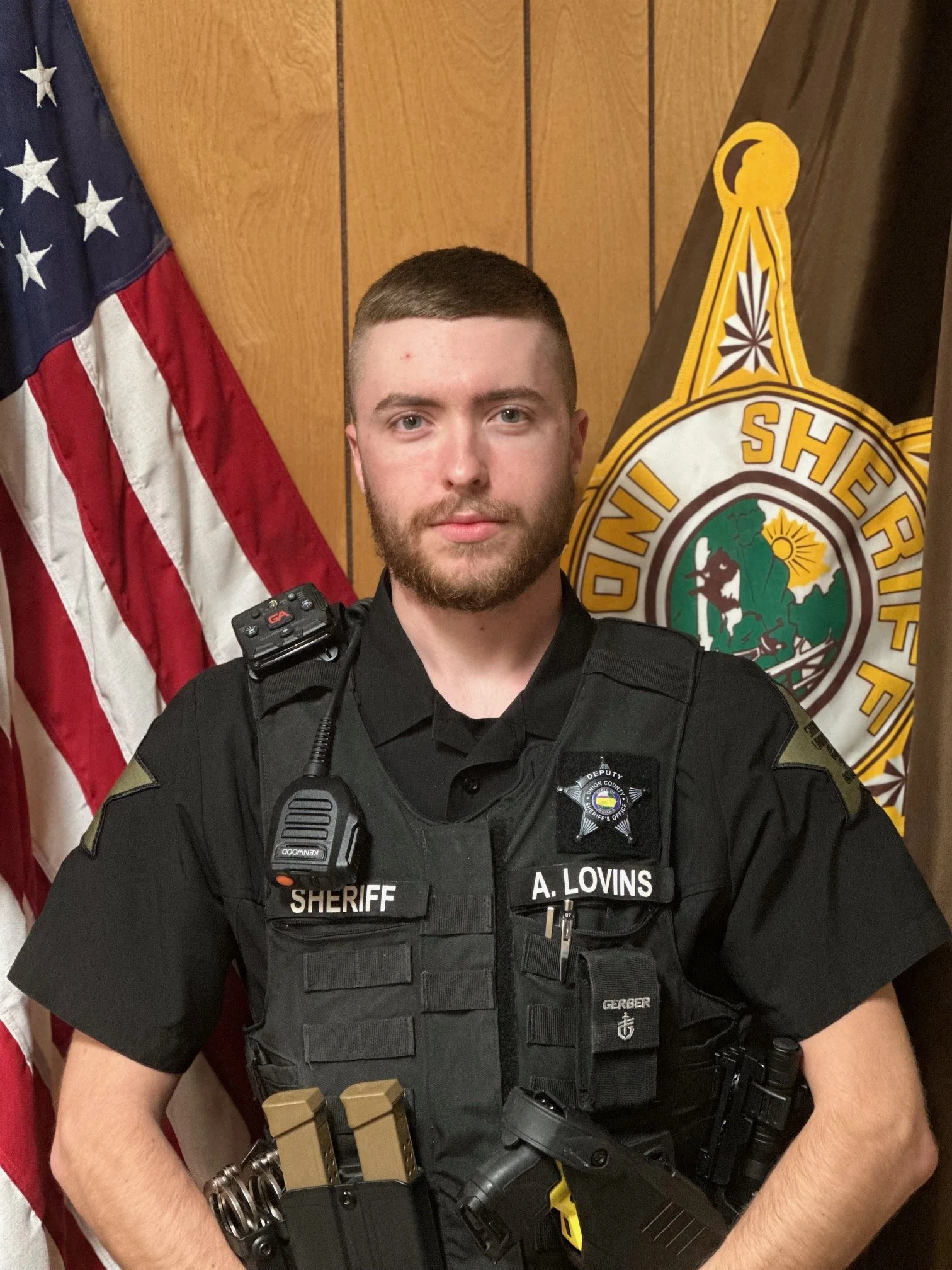 A male sheriff officer in uniform stands in front of an American flag and a sheriff badge insignia. He has a badge on his shirt and various equipment attached to his vest, including a radio, magazines, and a firearm.