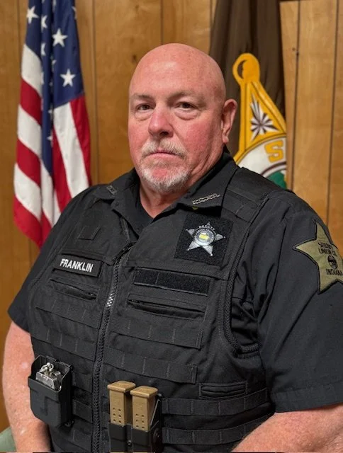 A police officer in uniform standing indoors with an American flag and a government flag in the background.