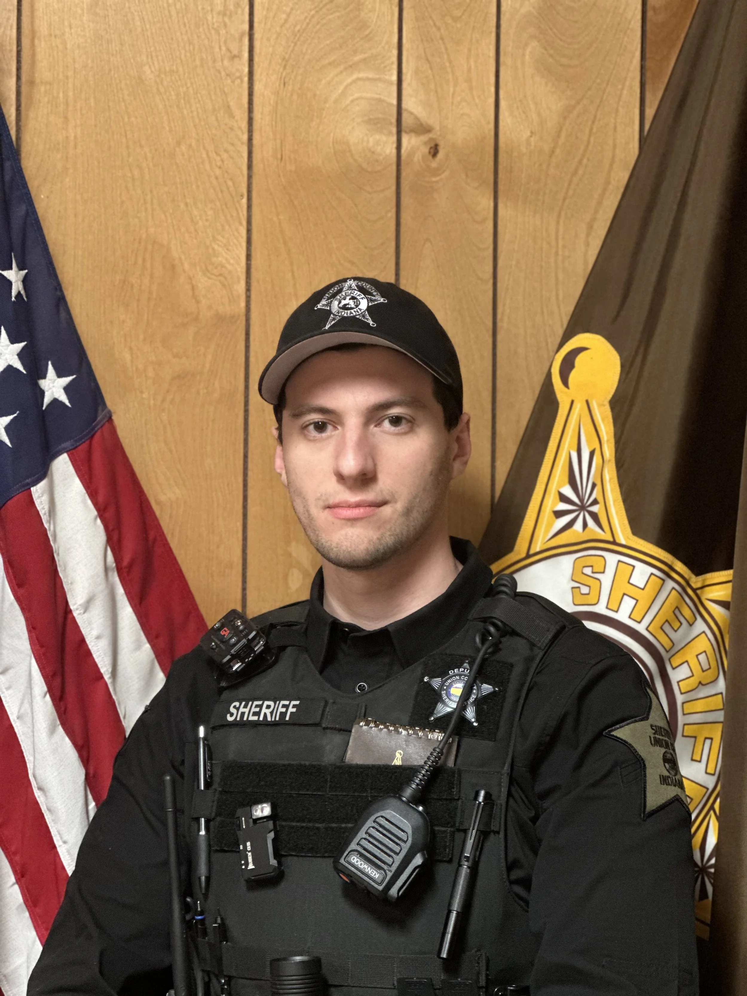 A male sheriff officer taking a selfie in front of an American flag and sheriff badge on a wooden wall.