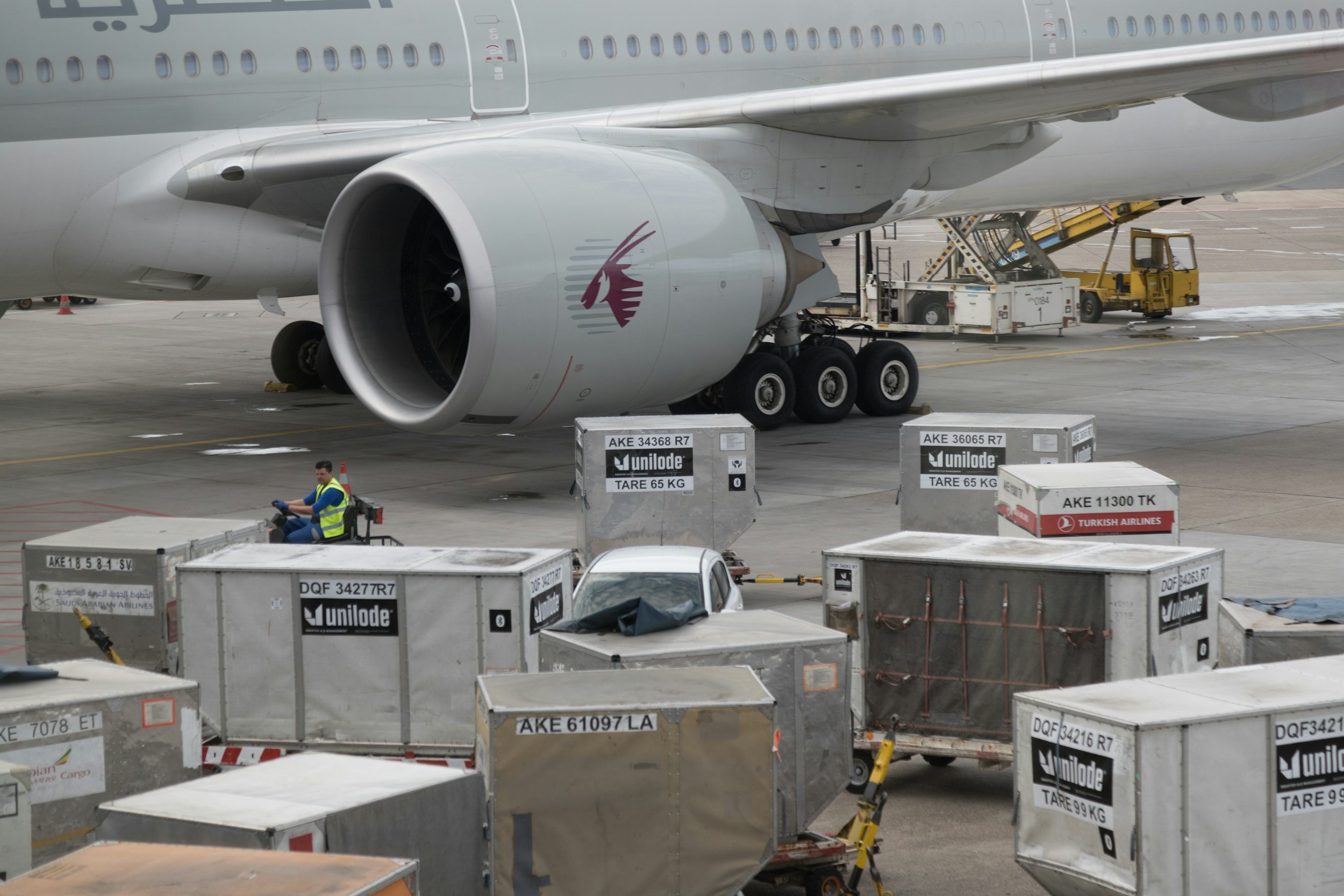 A close-up view of a Qatar Airlines airplane at an airport gate, with luggage carts and cargo containers nearby.