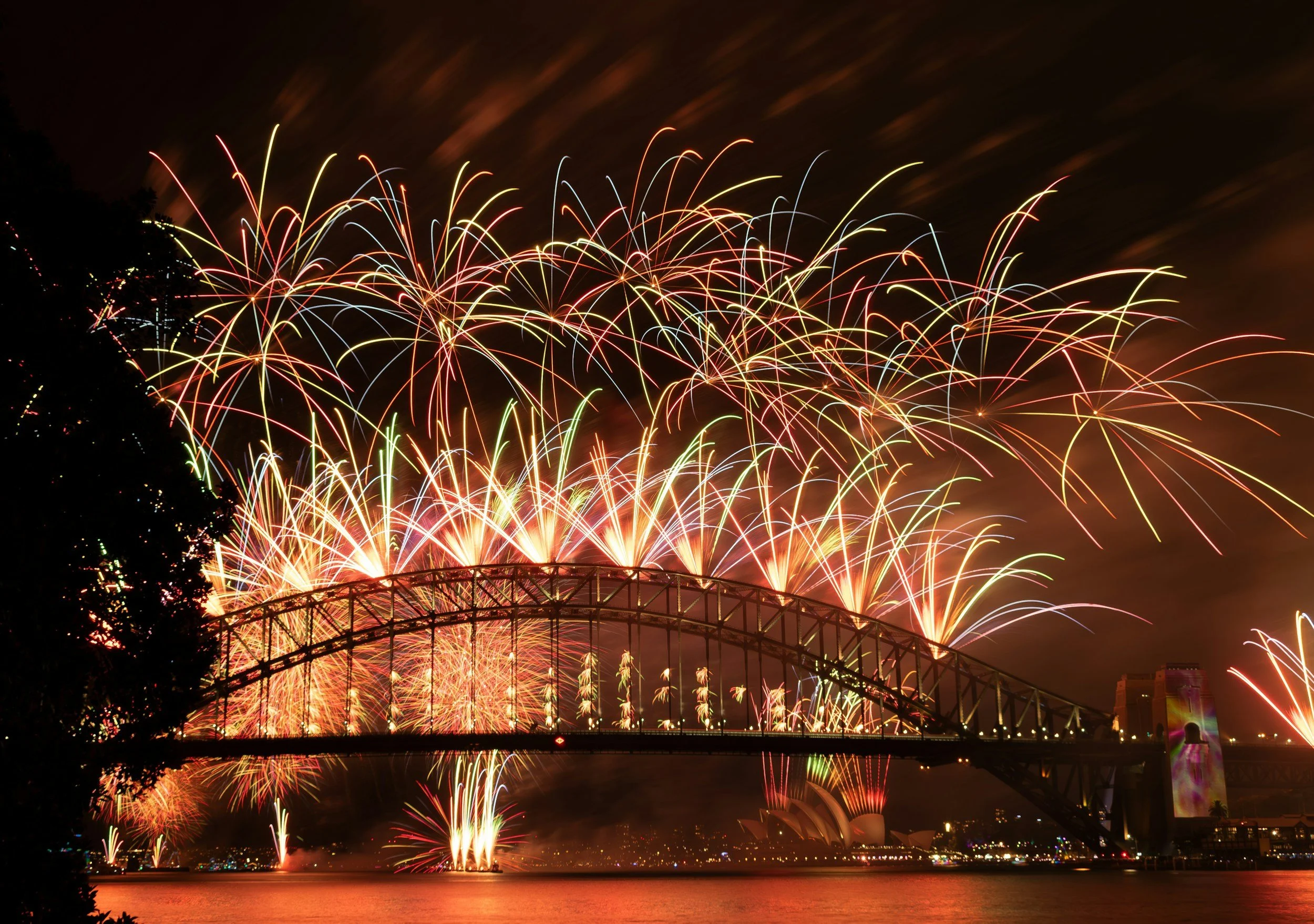 Fireworks display over a bridge at night, with reflections on the water below.