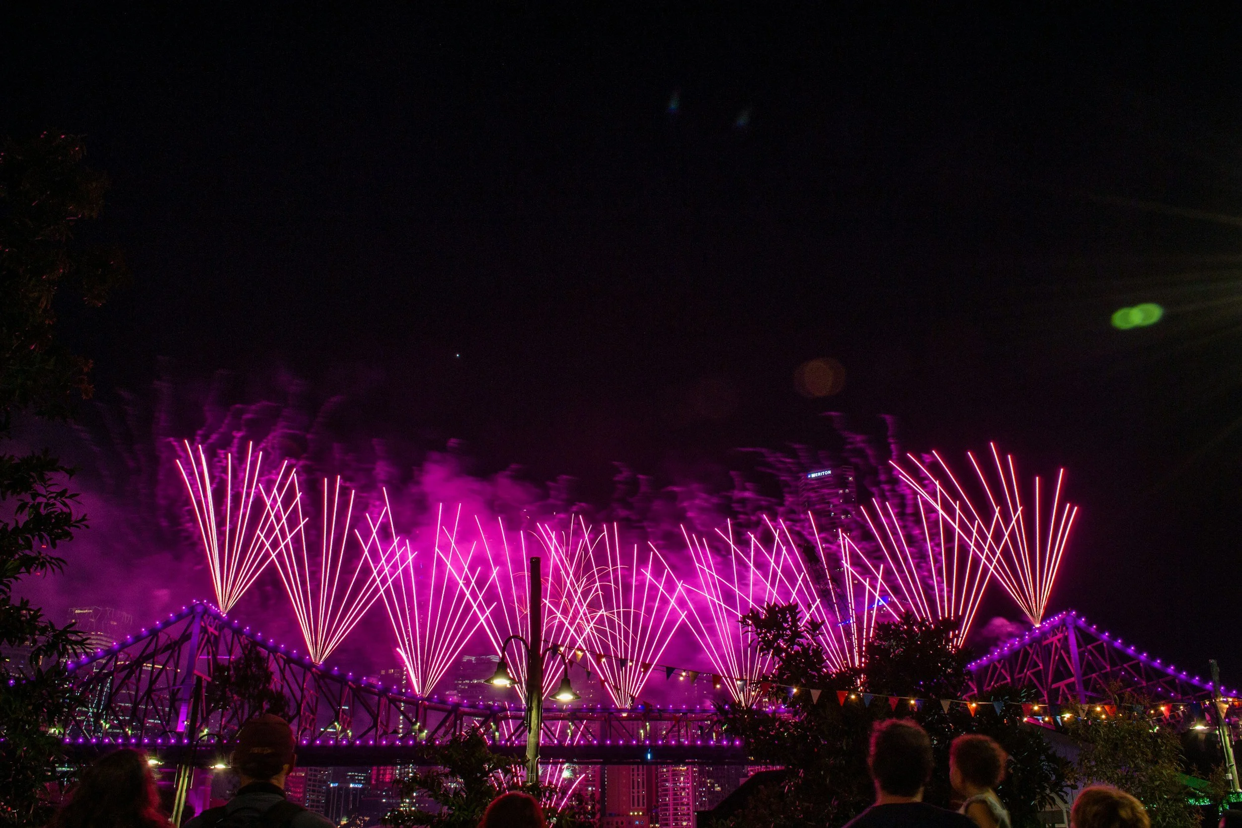 Nighttime scene of a fireworks display with pink and purple fireworks over a cityscape, with many people watching in the foreground.