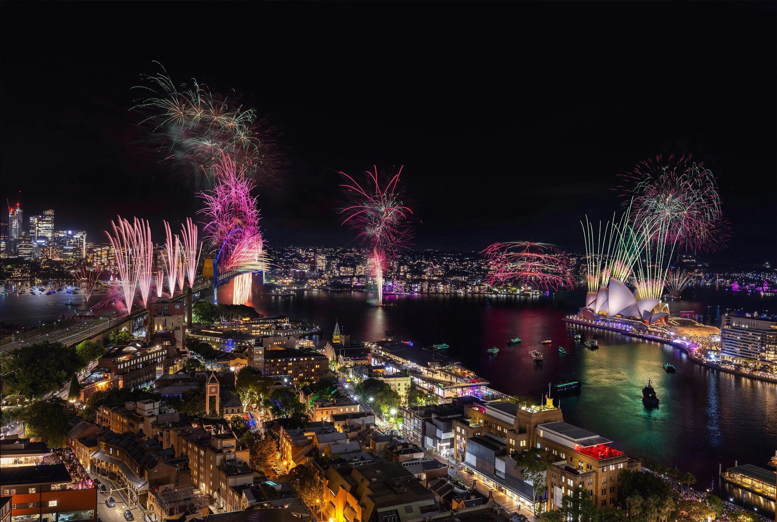 Nighttime cityscape with fireworks over the Sydney Opera House and Harbour Bridge, illuminated buildings, boats on the water, and streets with traffic.
