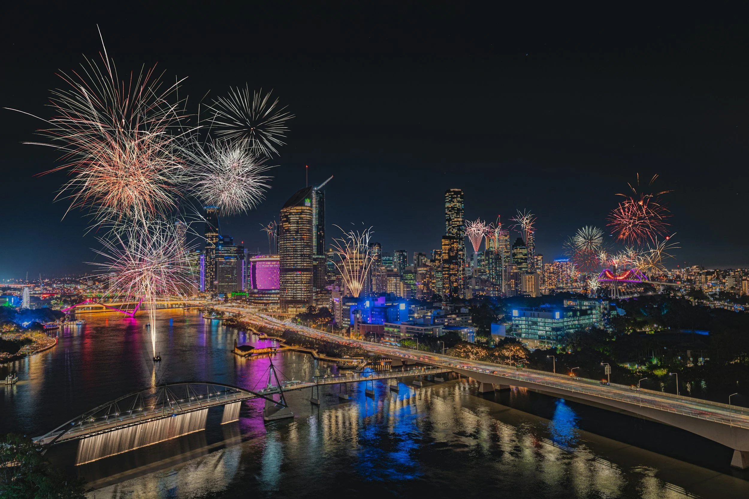 Nighttime cityscape with fireworks over the skyline and a river reflecting the lights.