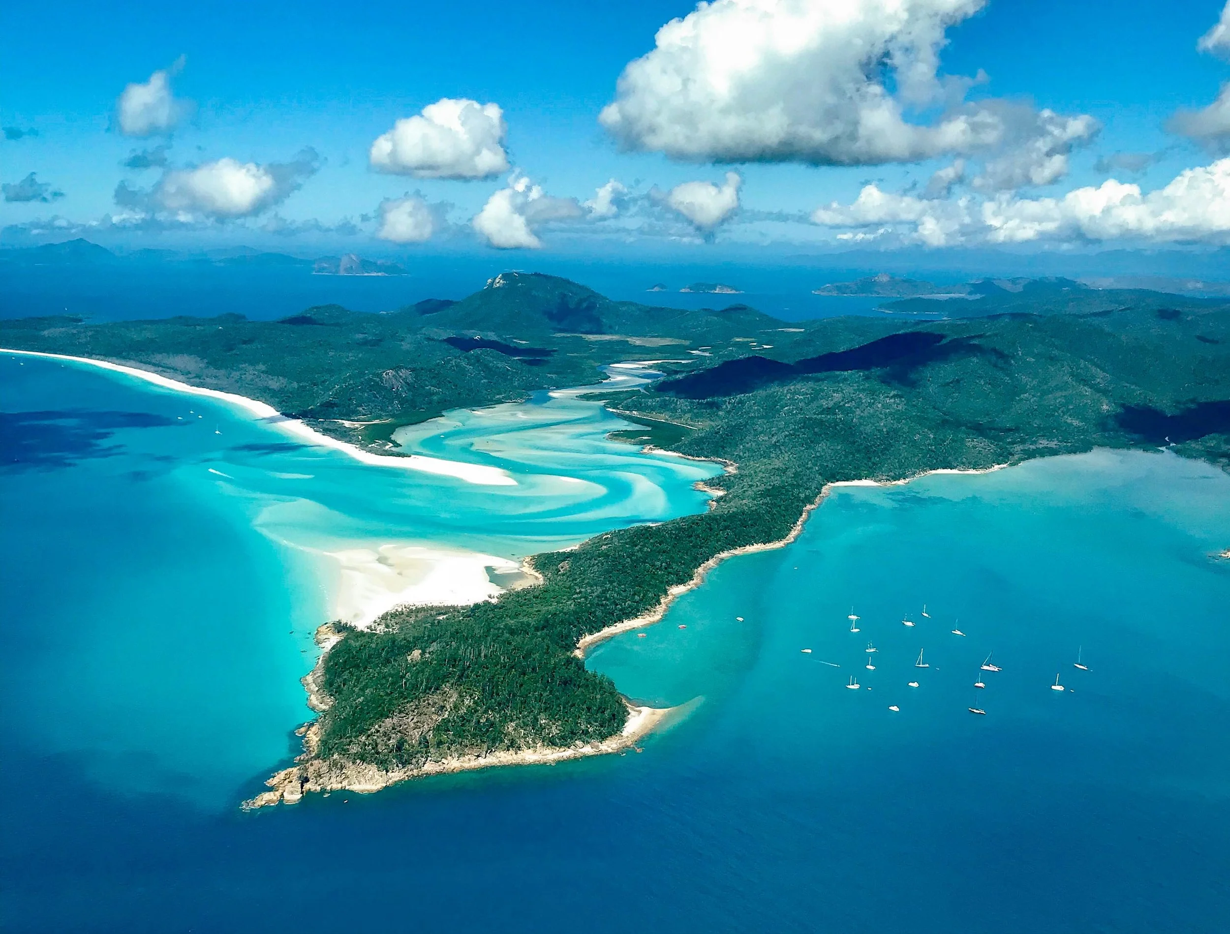 Aerial view of a coastal area with turquoise waters, sandy beaches, green islands, and sailboats anchored near the shore, under a partly cloudy sky.