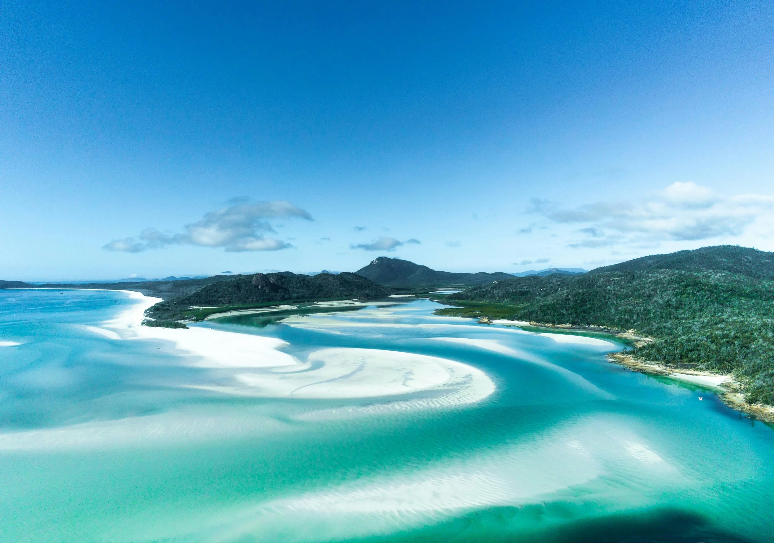 A landscape view of a coastal area with sandy beaches, turquoise water, green hills, and distant mountains under a partly cloudy sky.