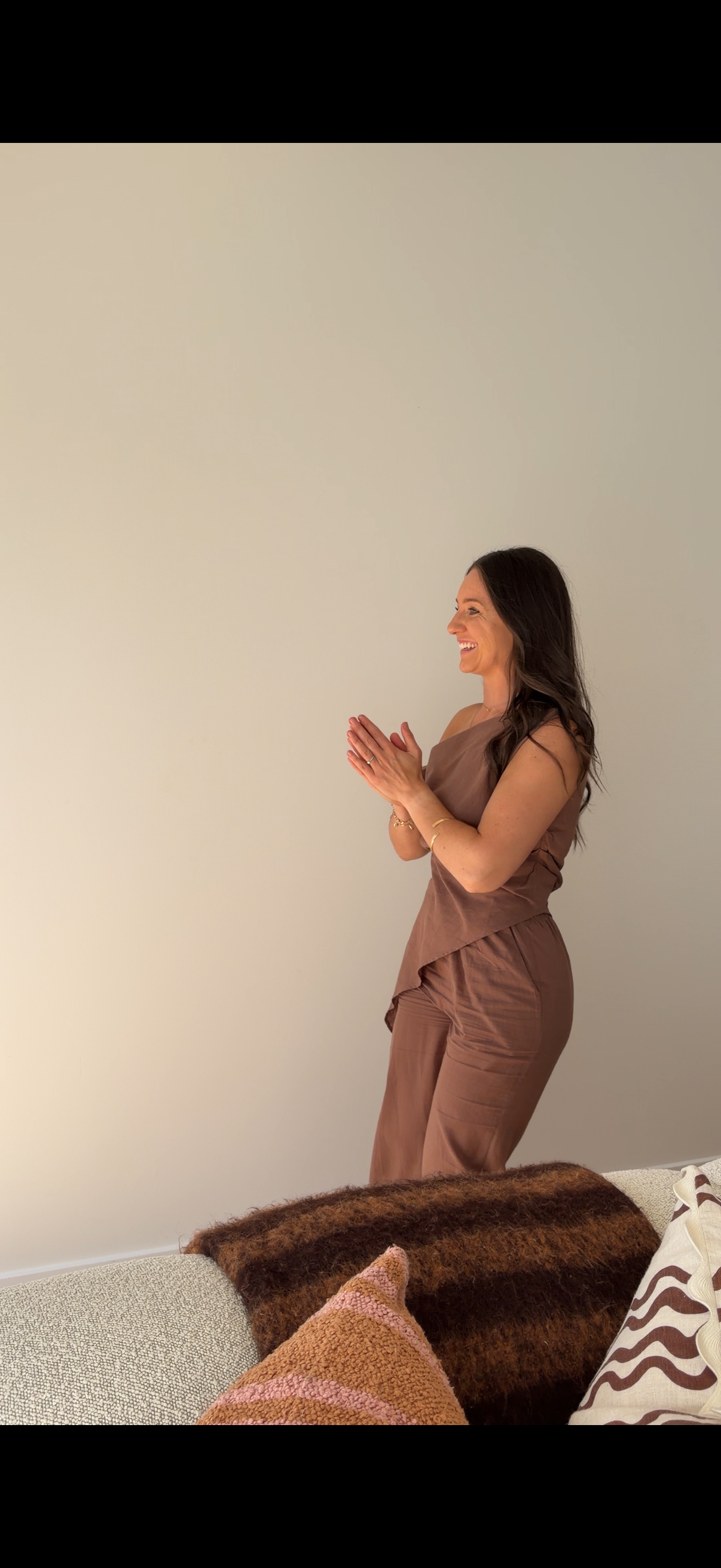A woman standing indoors with her hands together in a prayer gesture, smiling and looking to the right, against a plain white wall. She is wearing a brown sleeveless top and matching pants. There is a sofa with pillows in front of her.
