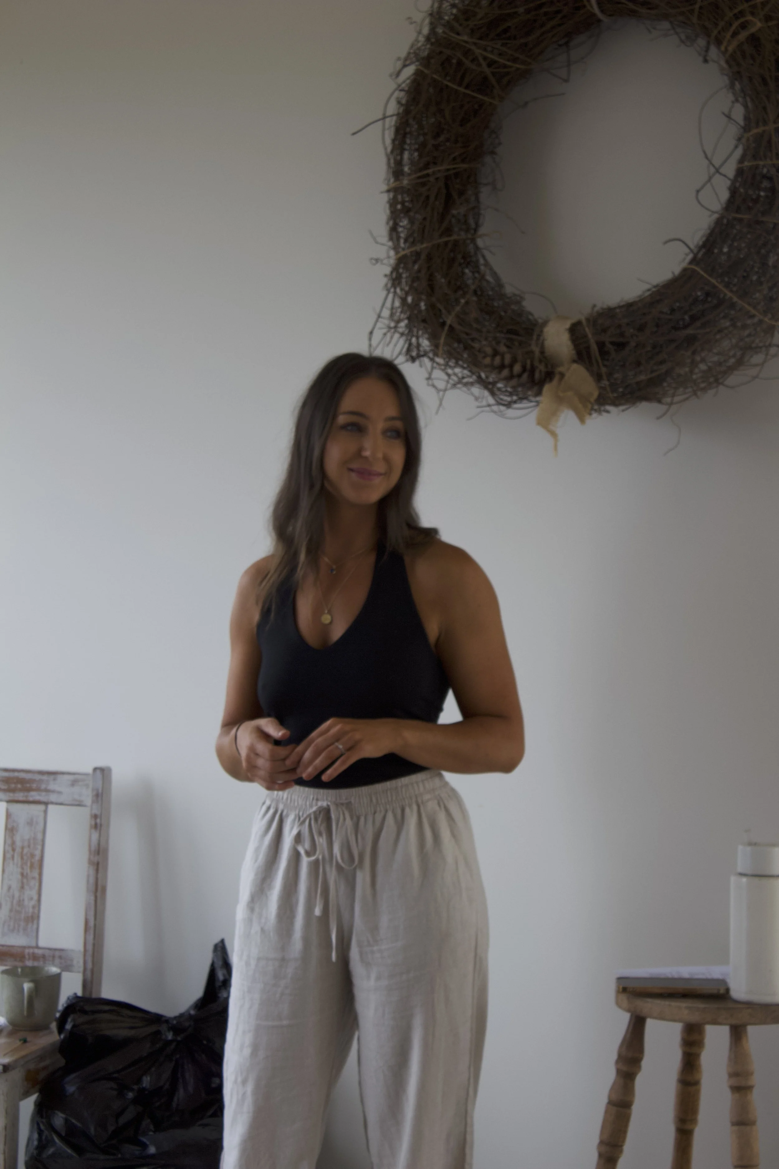 A woman with long brown hair wearing a black tank top and beige drawstring pants, standing indoors against a plain white wall with a decorative wreath hanging above her head. There are some furniture and bags in the background.