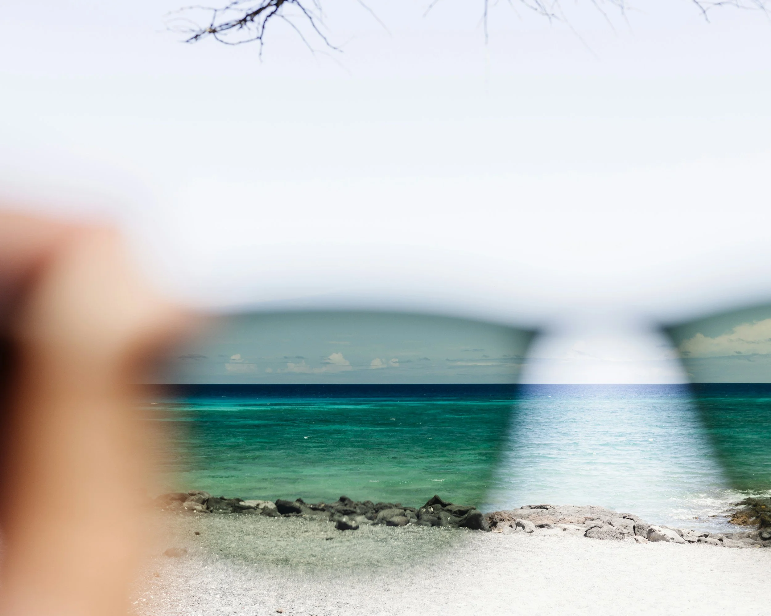 A scenic beach view with clear turquoise water, rocks on the shore, and leafless tree branches overhead.