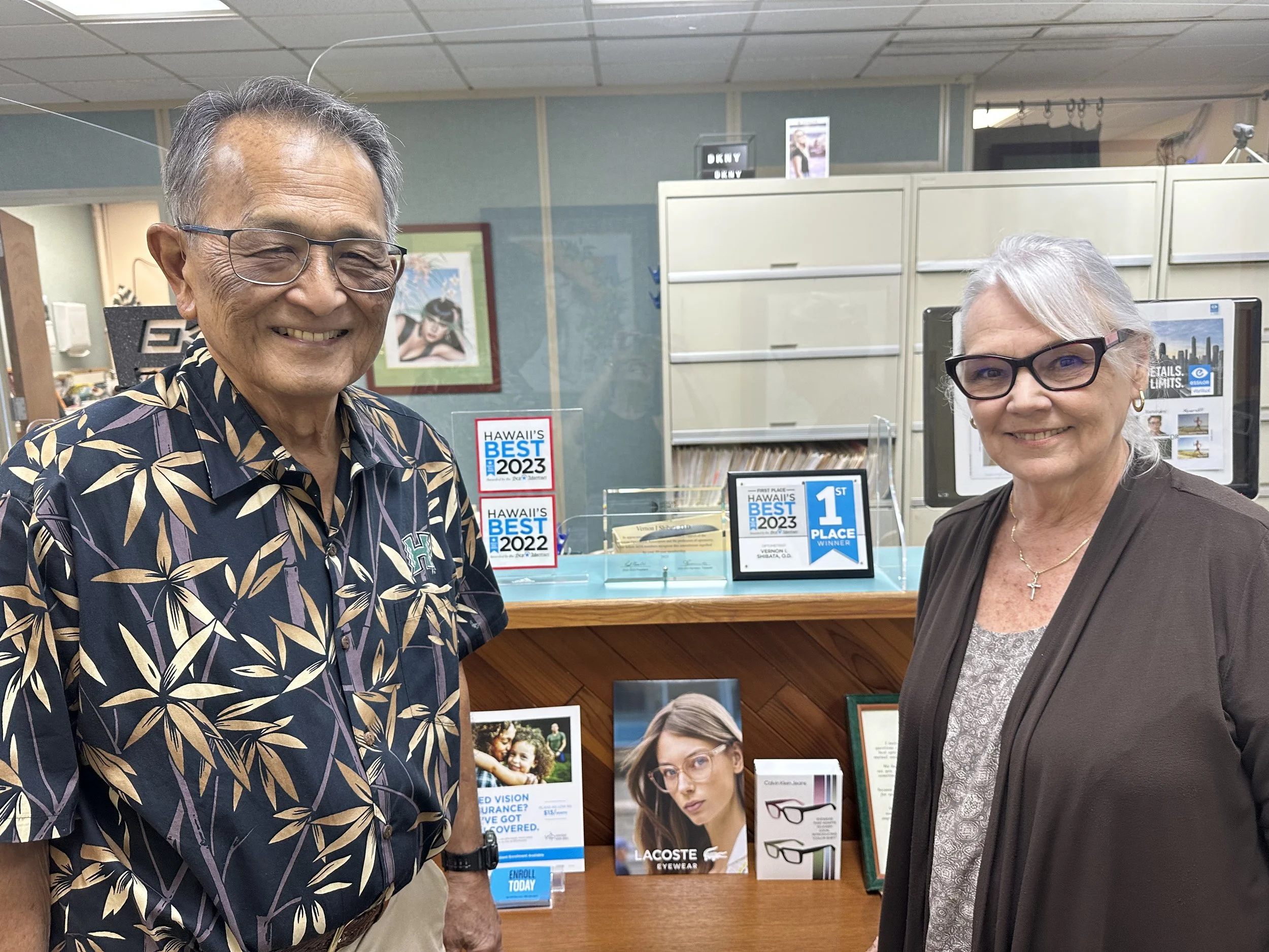 Dr. Shibata and Mrs. Shibata standing and smiling with their award featuring "Star Advertiser's Hawaii's Best 2023, 1st Place" for Optometrists in Honolulu. There are framed pictures, brochures, and eyewear advertisements visible behind them.