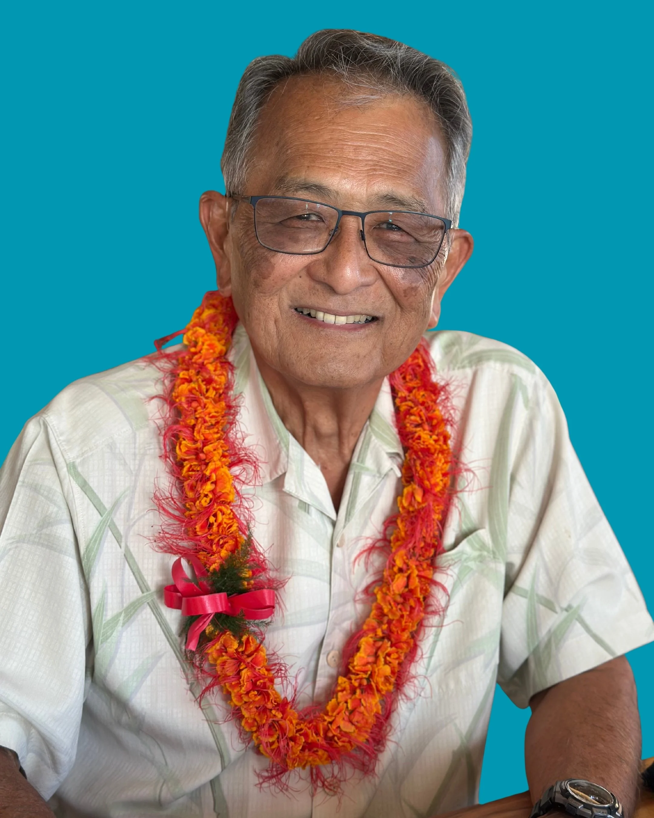 An elderly man wearing glasses and a light-colored, patterned shirt, smiling and adorned with a garland of orange flowers with red accents and a red ribbon, set against a teal background.