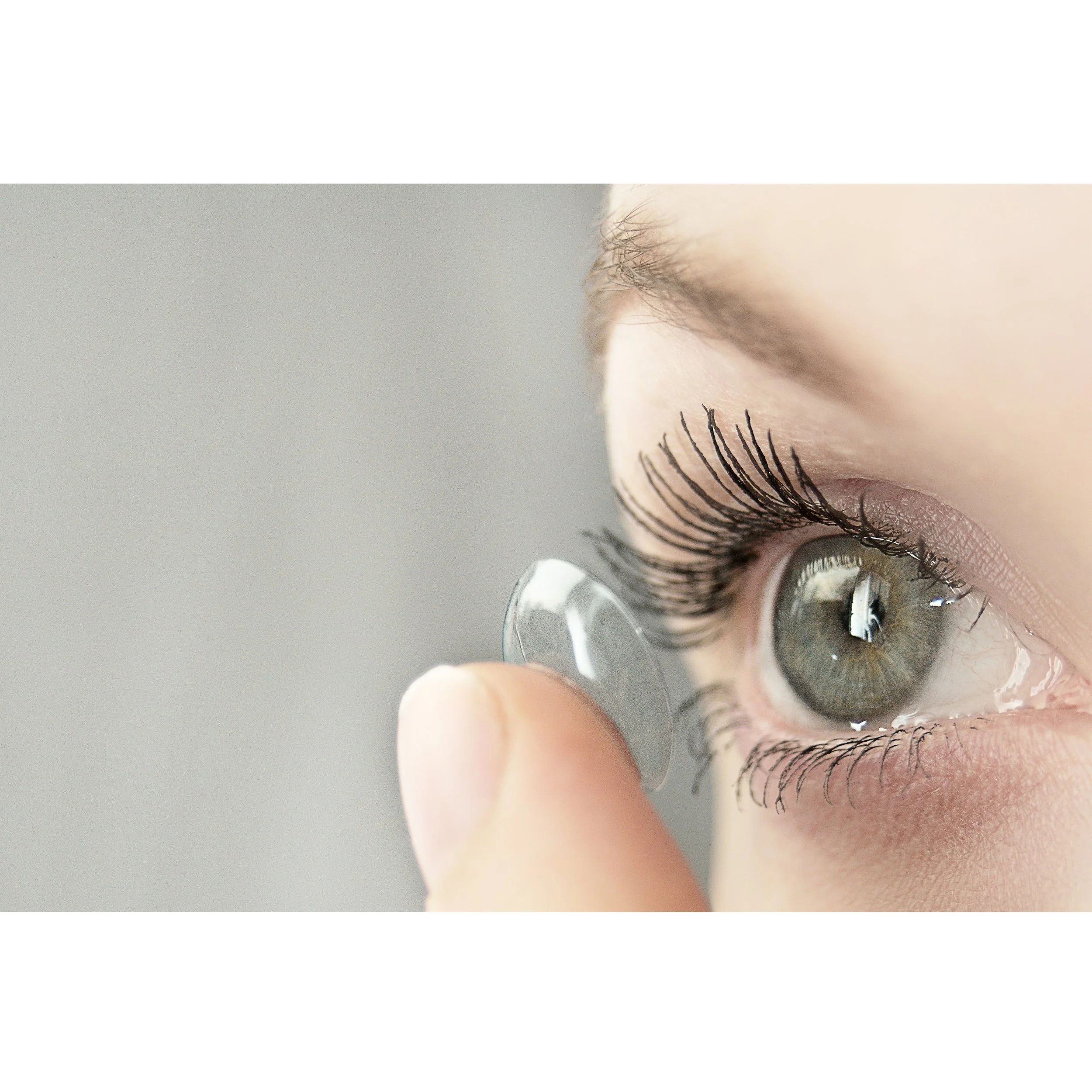 Close-up of a person’s eye with long eyelashes and a contact lens, with a finger holding a contact lens at the lower edge of the eye.