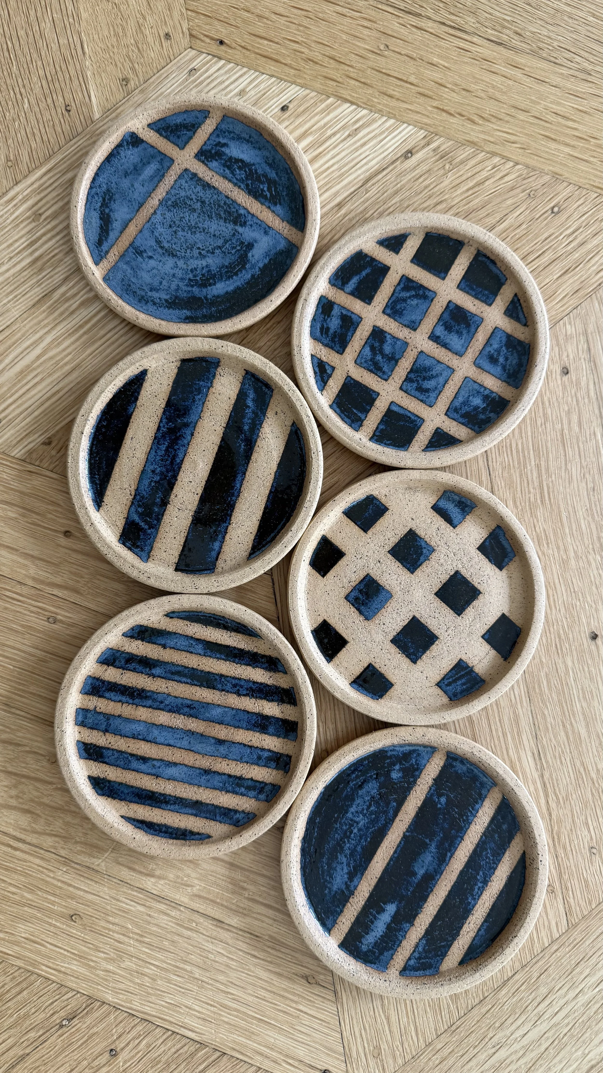 Six small round ceramic coasters with blue painted geometric patterns on tan backgrounds, arranged on a wooden surface.