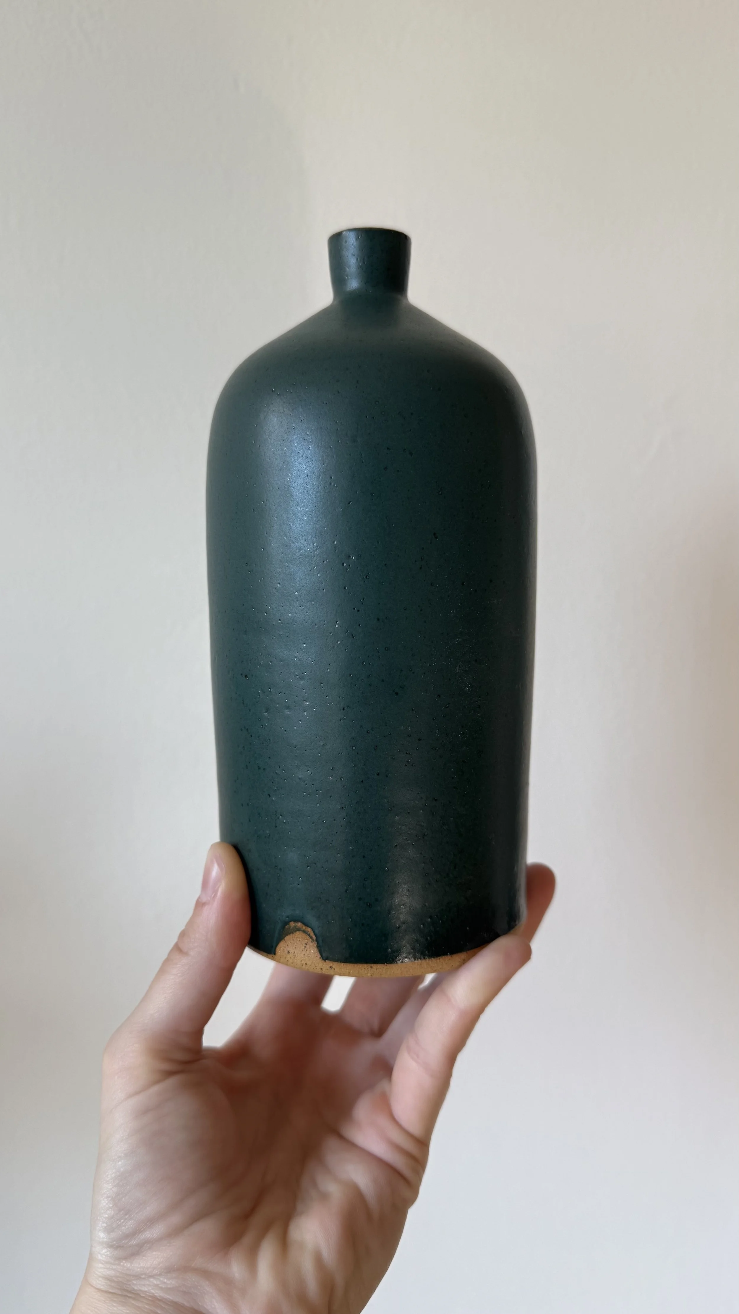Hand holding a dark green ceramic vase with a small opening, against a plain light-colored wall.