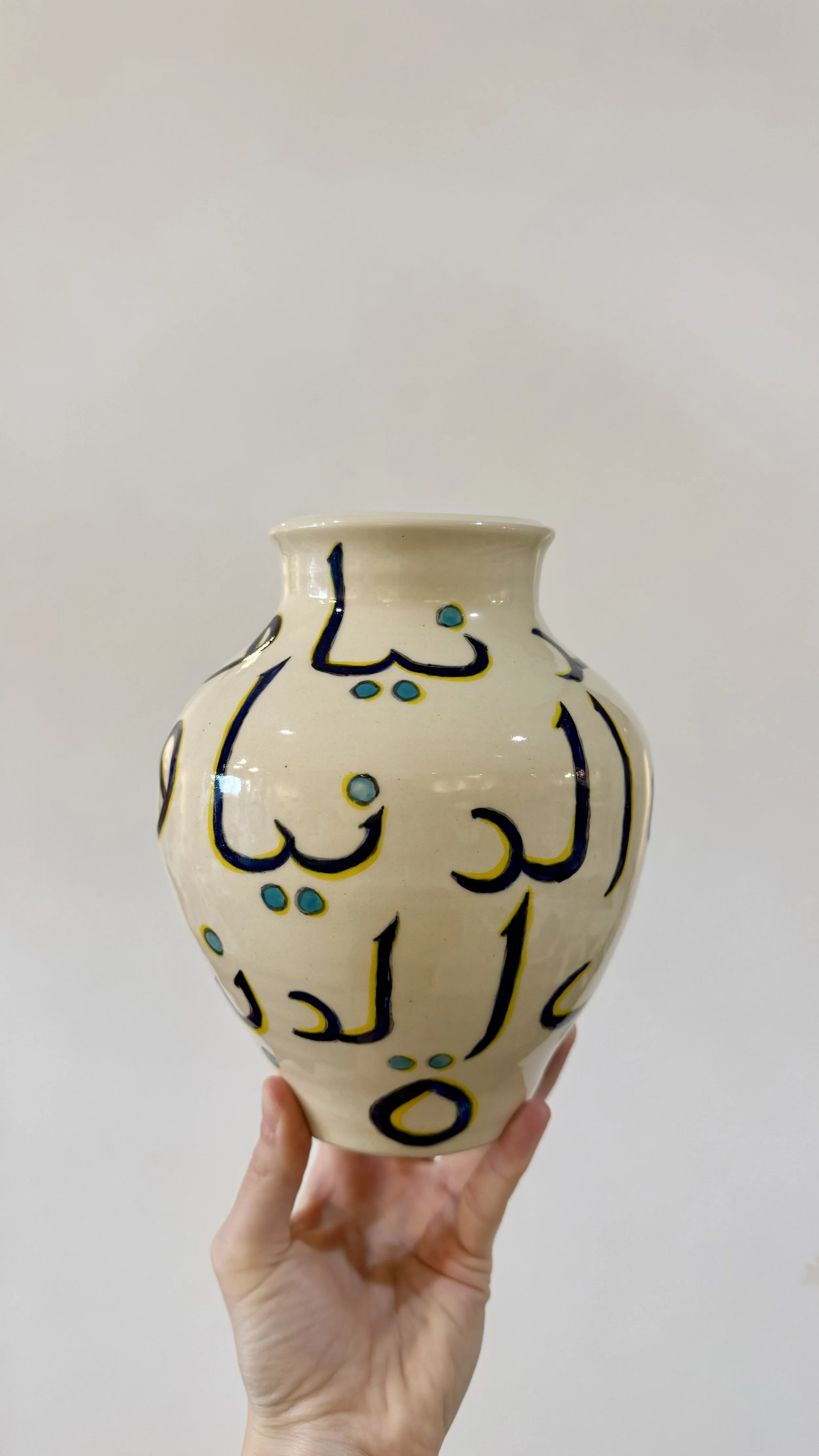 A hand holding a ceramic vase with black and blue Arabic calligraphy, against a plain off-white background.