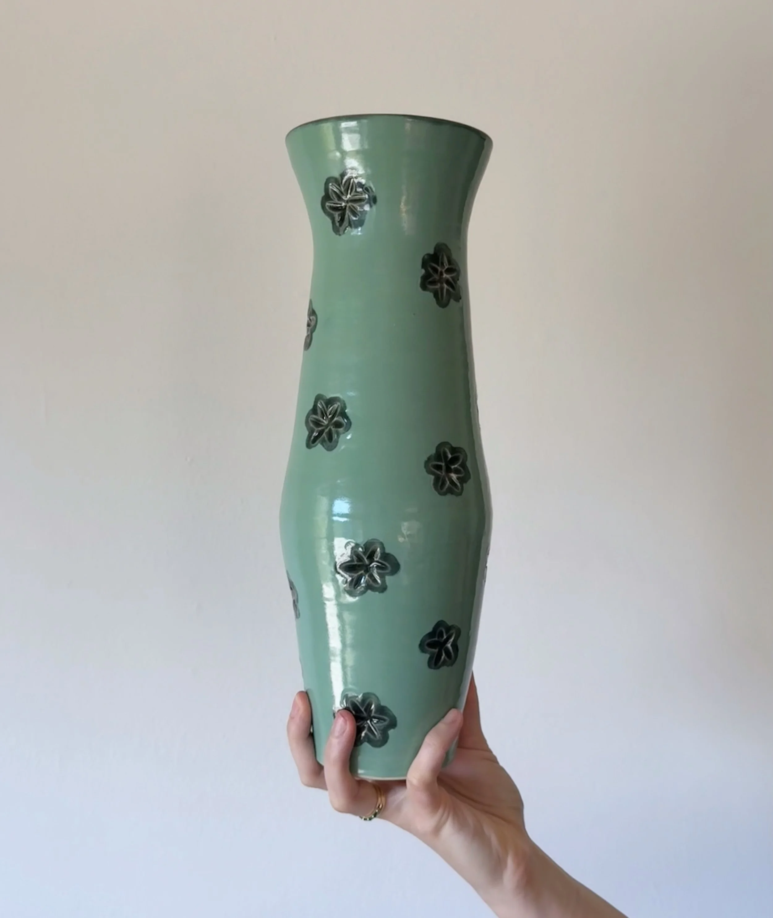 A person is holding a tall light green ceramic vase decorated with dark green floral designs against a plain white wall background.